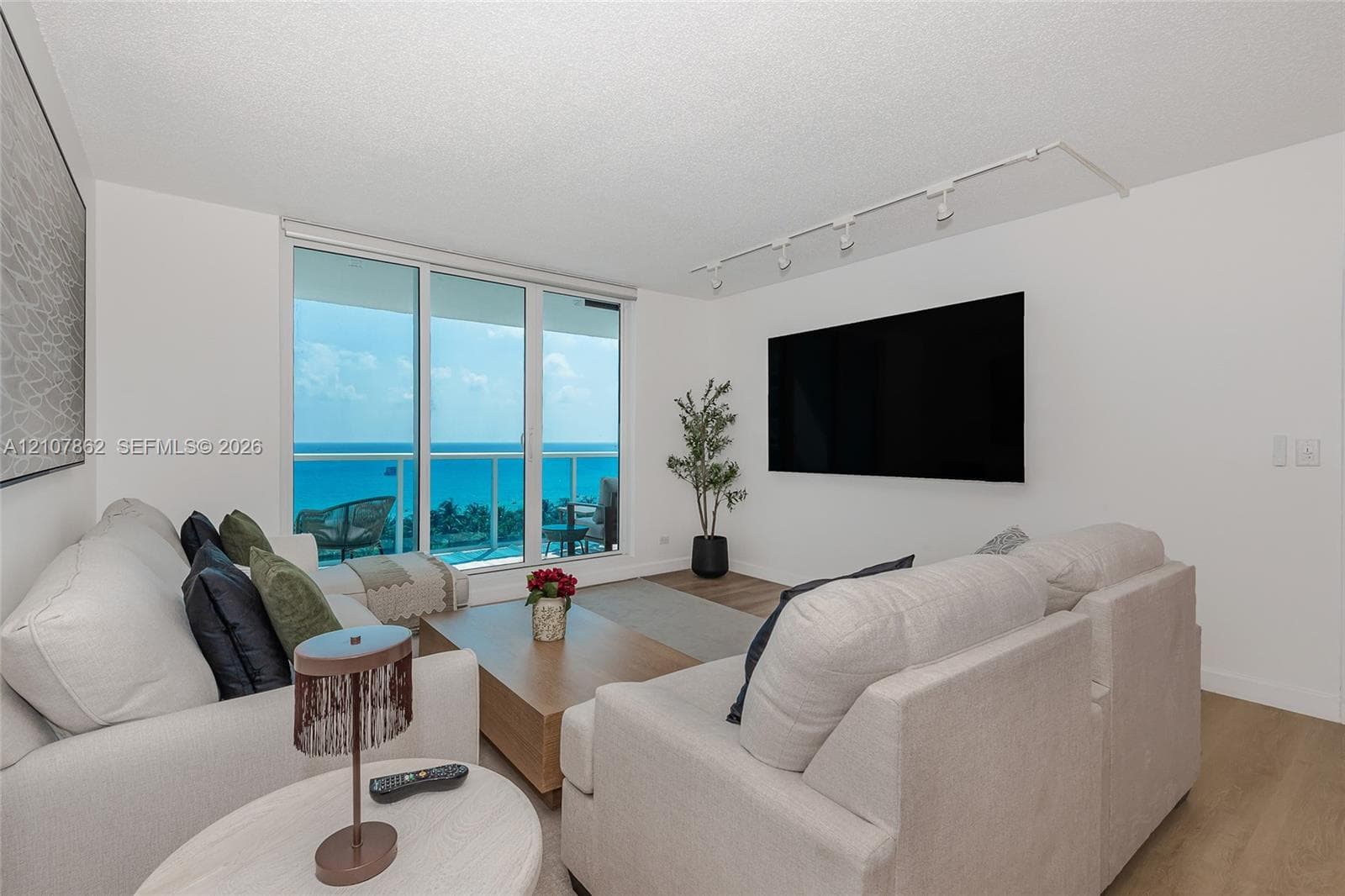 Floor-to-ceiling glass sliding doors, wood-finish flooring, adjustable track lighting, and a private balcony featuring an ocean backdrop