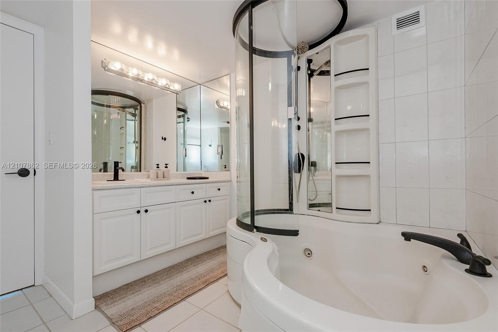 Bathroom featuring a white vanity cabinet with a light-toned countertop, an integrated jetted tub with a curved glass-enclosed shower, and a full-width wall mirror with integrated vanity lighting