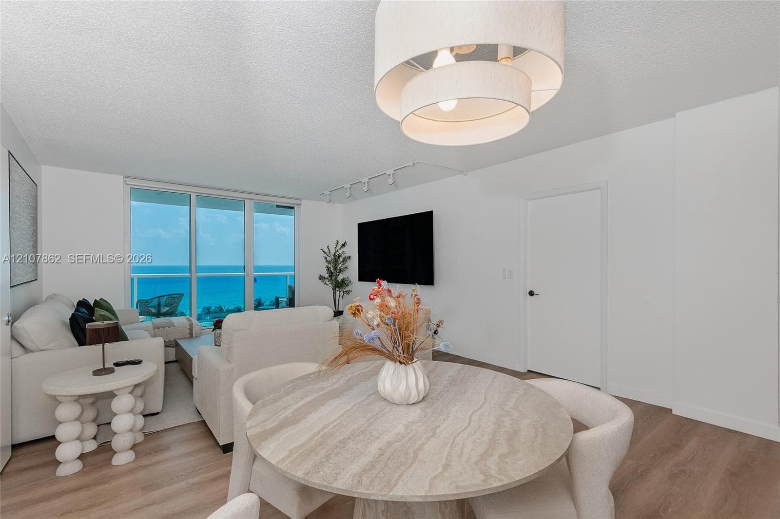 Open-concept interior featuring wood-finish flooring, a contemporary multi-tiered drum chandelier, recessed track lighting, and floor-to-ceiling glass panel doors revealing ocean scenery