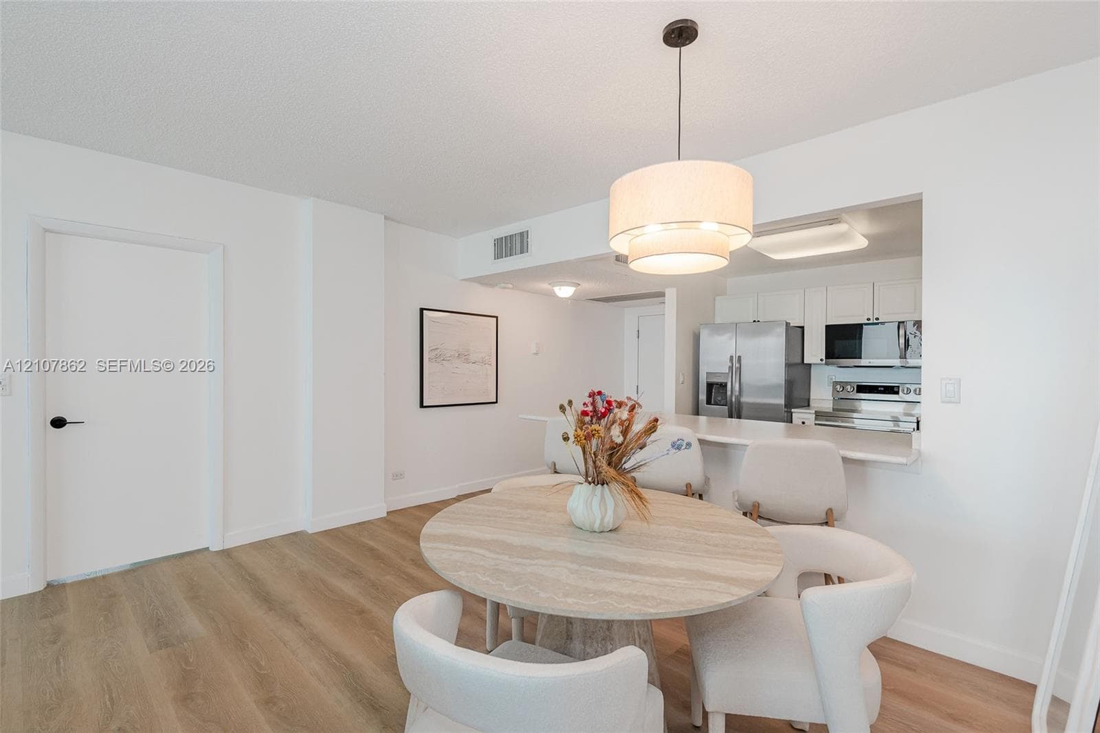 Dining area featuring wood-finish flooring, a tiered drum pendant light, and a pass-through opening to the kitchen equipped with stainless steel appliances and white cabinetry