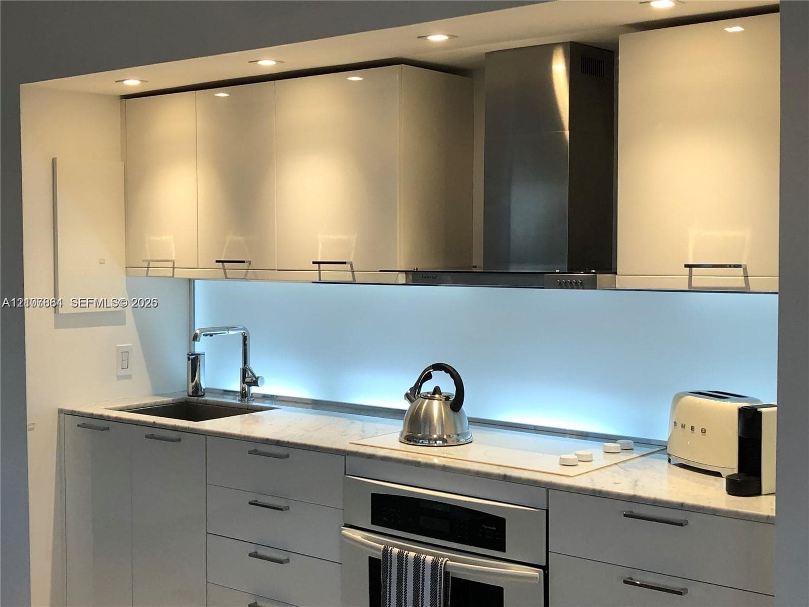 Kitchen featuring high-gloss cabinetry, stone countertops, stainless steel range hood, built-in cooktop, and under-cabinet lighting