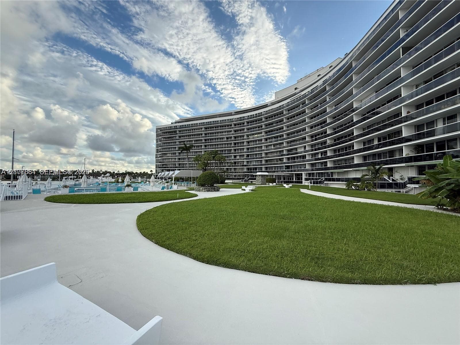 Curved mid-rise architectural facade featuring expansive wraparound balconies, manicured lawn areas, paved walkways, and dockside waterfront access