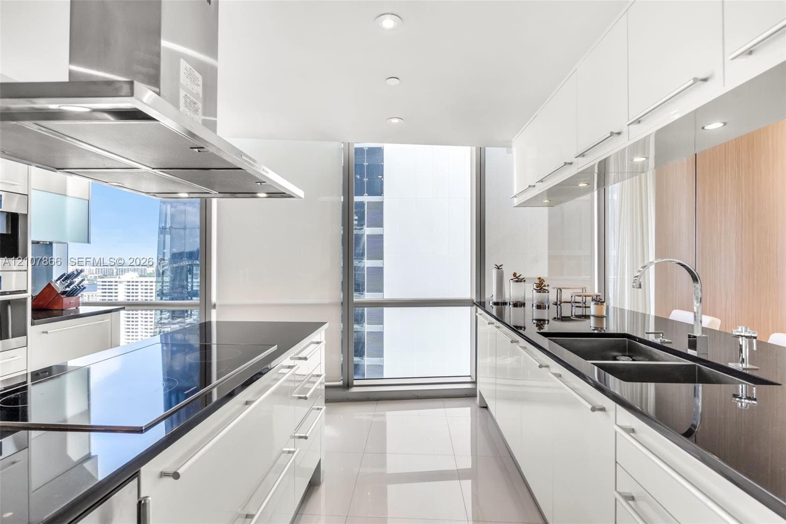 Contemporary kitchen featuring flat-panel white cabinetry, contrasting black stone countertops, an integrated stainless steel range hood, double under-mount sinks with a gooseneck faucet, and floor-to-ceiling glass windows