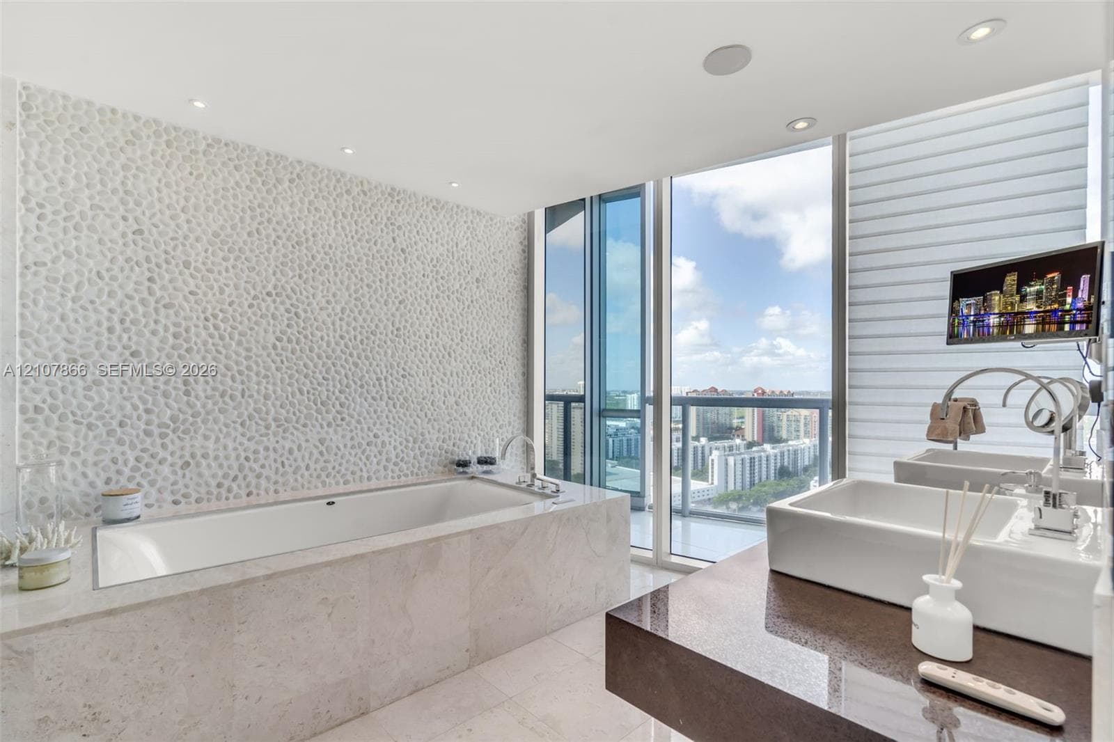 Drop-in bathtub with stone-finish surround, textured pebble-style accent wall, dual rectangular vessel sinks, polished stone countertop, and floor-to-ceiling windows