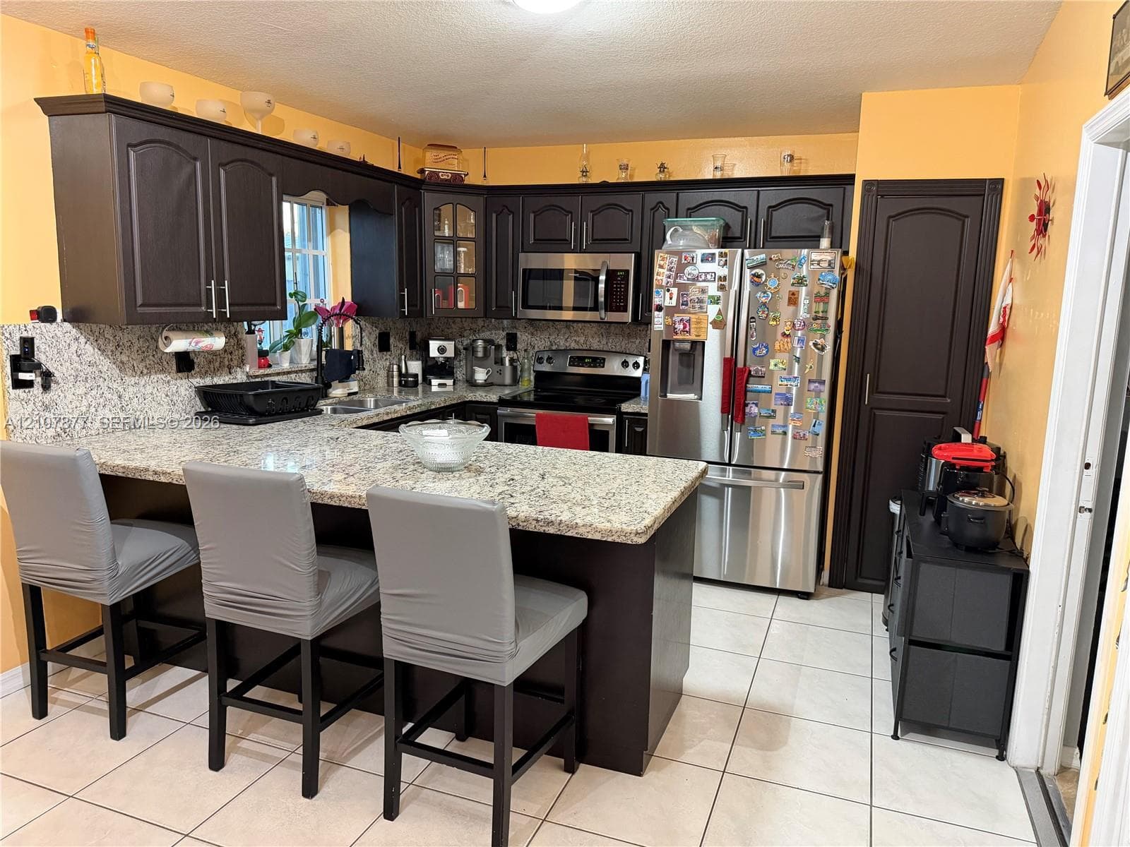 Kitchen featuring dark-toned cabinetry, light-colored stone-finish countertops, ceramic tile flooring, and integrated stainless steel appliances