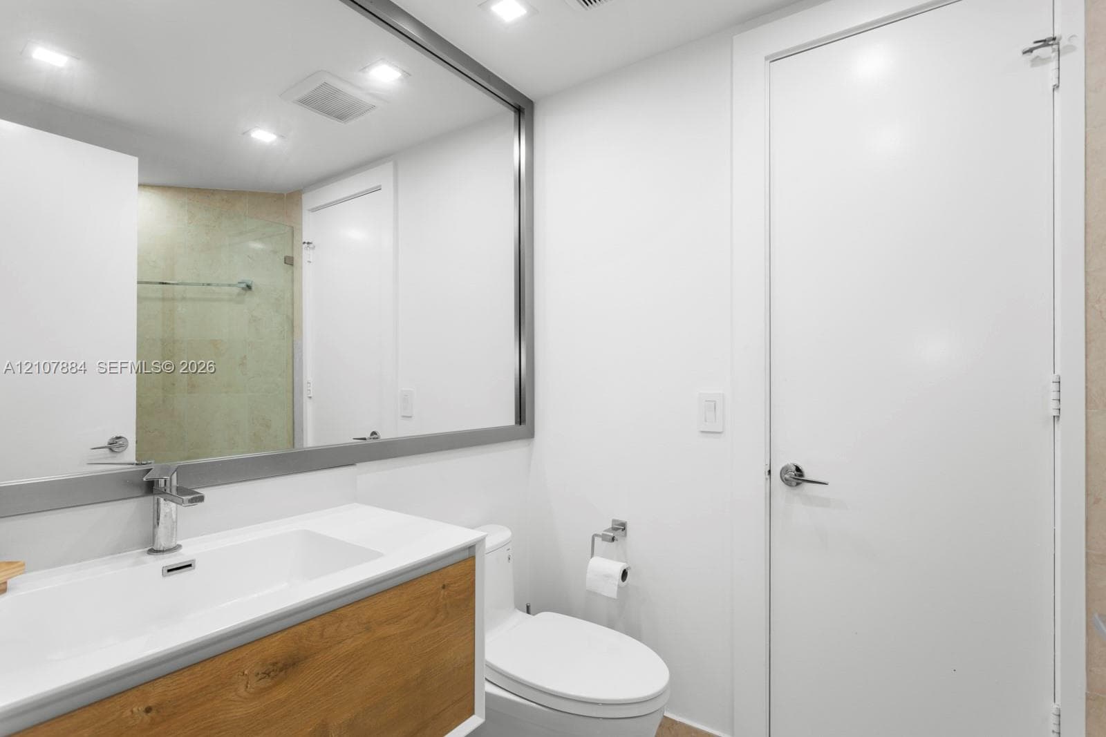 Floating vanity with a wood-finish cabinet and integrated rectangular sink, large wall-mounted mirror with a metallic frame, recessed ceiling lighting, and a glass-enclosed shower