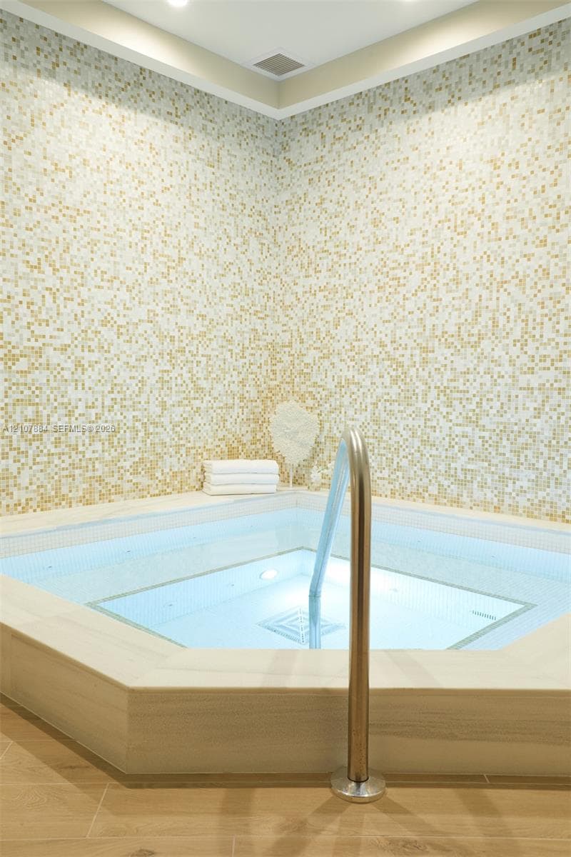 Built-in soaking tub featuring gold mosaic wall tile, stone-finish surround, wood-finish flooring, and a metallic handrail