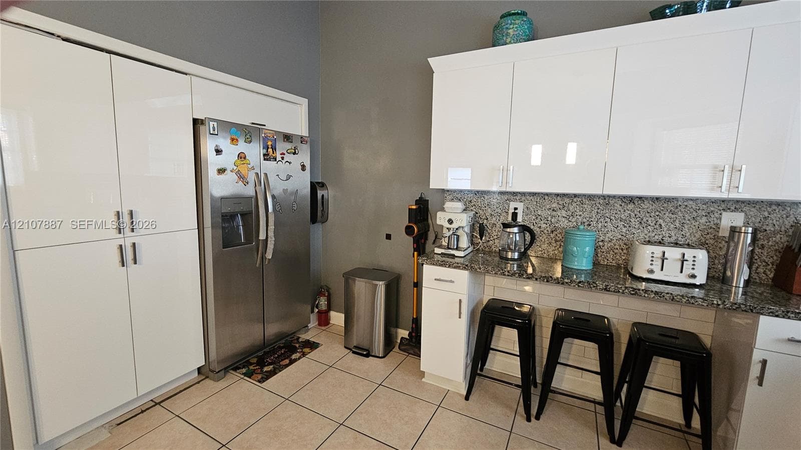 Kitchen featuring white high-gloss cabinetry, speckled stone countertops, a stainless steel side-by-side refrigerator, subway tile backsplash, and tile flooring