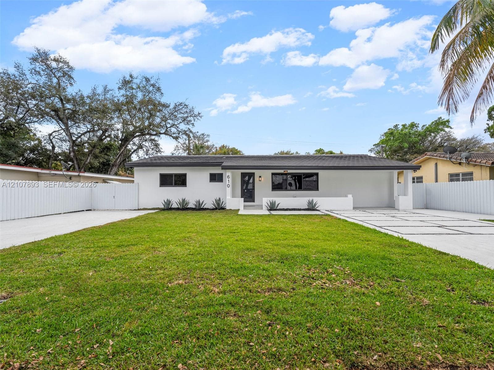 Single-story exterior featuring white stucco siding, a low-slope roof, expansive front lawn, concrete paver driveway with integrated turf, and full-perimeter privacy fencing