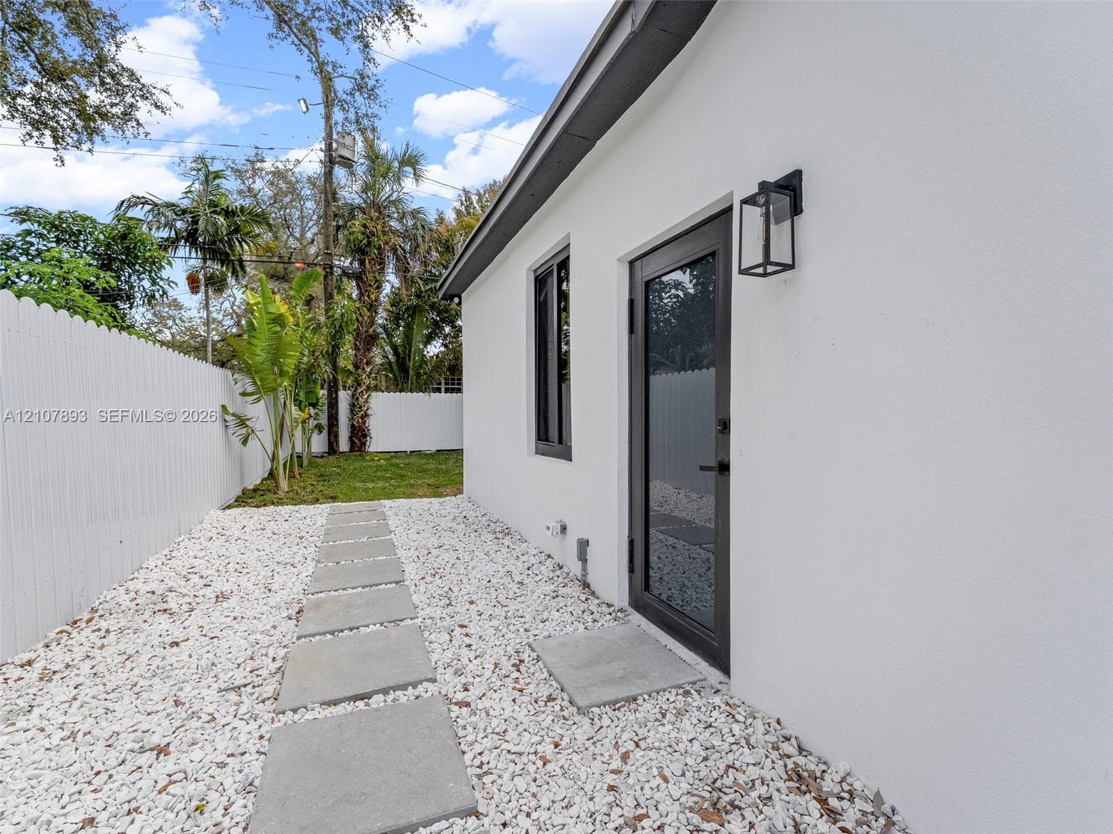 Stucco exterior featuring a dark-framed glass door, exterior wall-mounted lantern lighting, white privacy fencing, and a stone-paver walkway set in gravel
