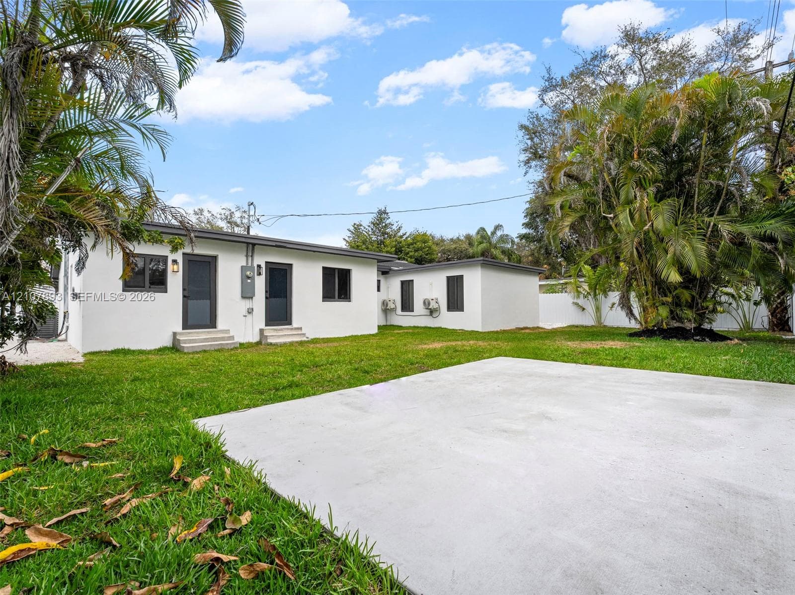 Expansive backyard featuring a concrete patio, white exterior facade, dark-trimmed windows, and mature palm trees