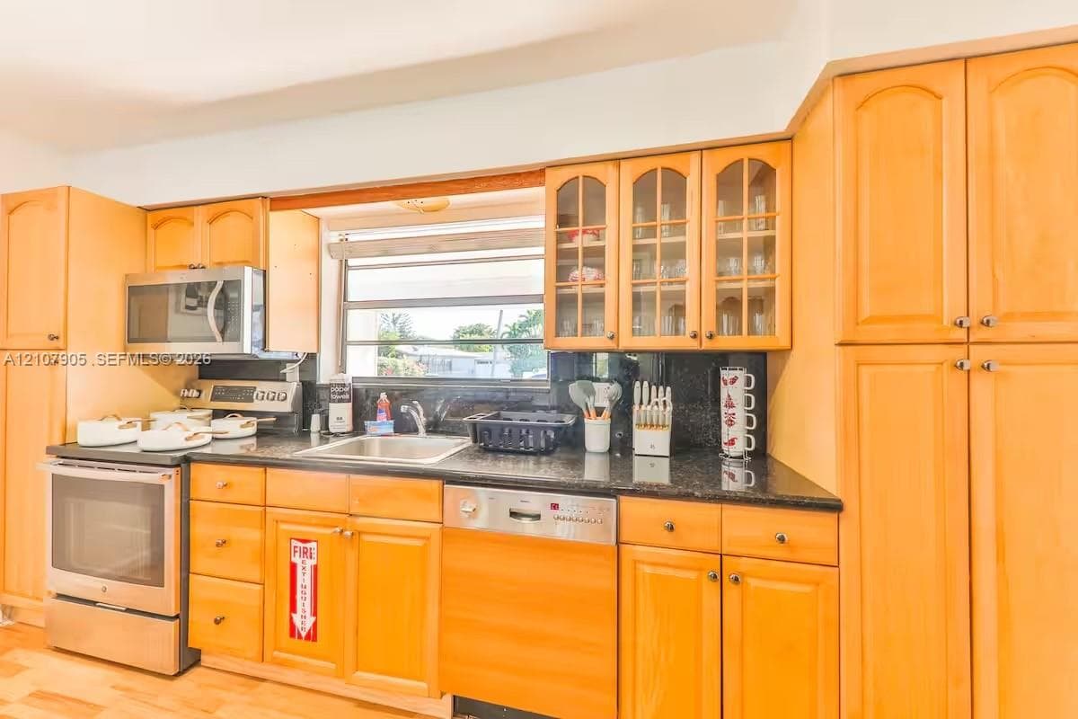 Kitchen featuring natural wood cabinetry, stone countertops, stainless steel appliances, glass-panel upper cabinet doors, and a window above the sink