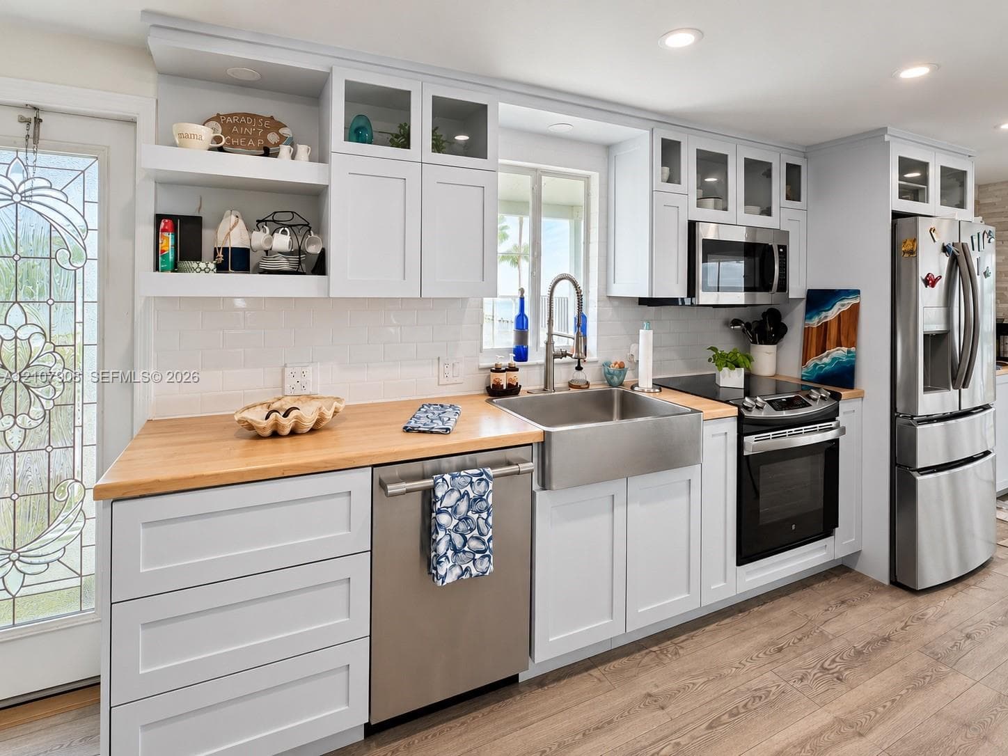 Kitchen featuring white shaker-style cabinetry, wood-finish countertops, stainless steel farmhouse sink, glass-front upper cabinet doors, and wood-finish flooring