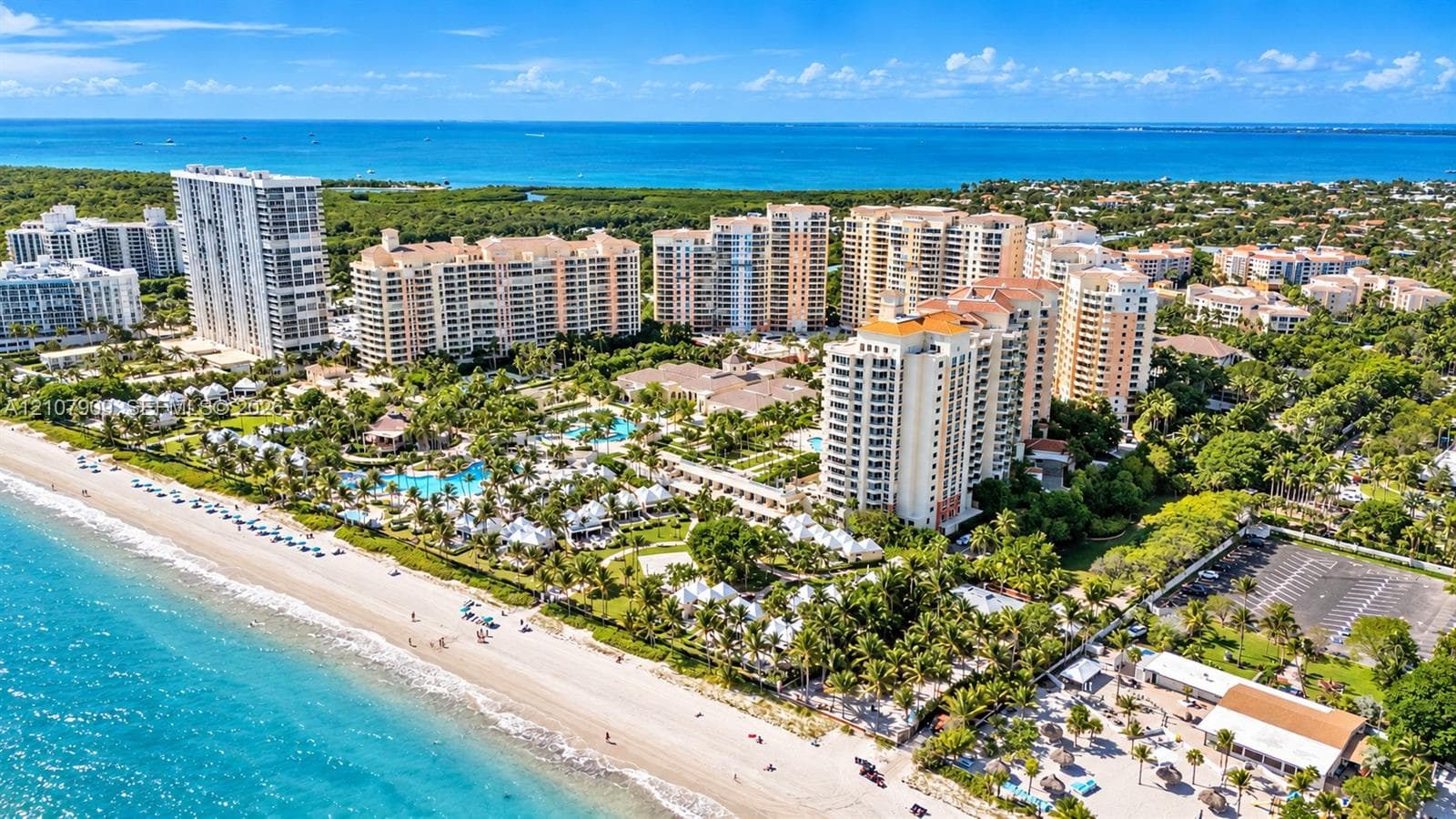 Aerial perspective of oceanfront high-rise structures, featuring expansive sandy coastline, turquoise water, manicured tropical landscaping, and outdoor swimming pools