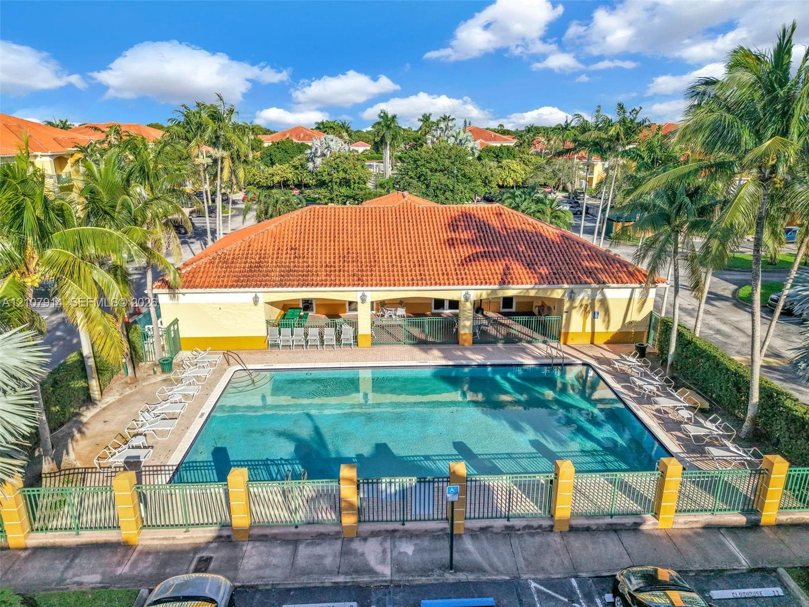 Community swimming pool featuring a rectangular basin, perimeter metal fencing, masonry pillars, and a covered pavilion with a terracotta tile roof, surrounded by mature palm trees