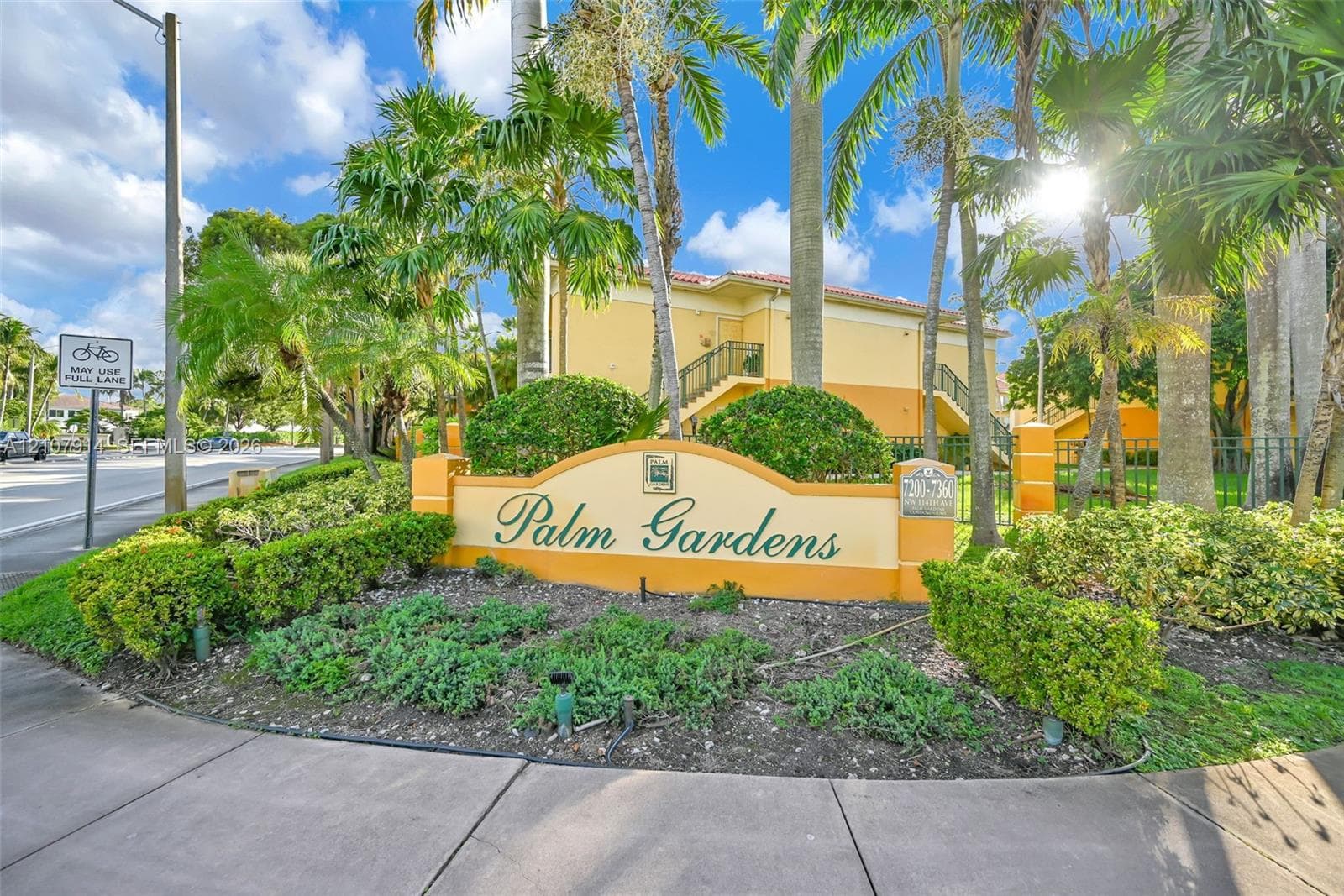 Stucco exterior community signage featuring masonry pillar accents, decorative landscaping, and mature palm trees