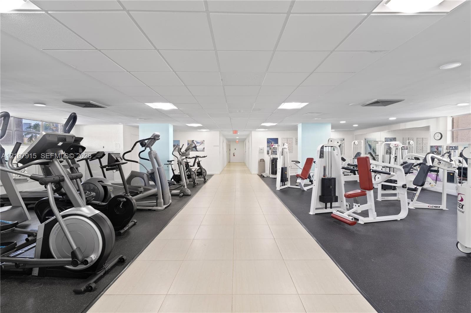 Fitness center featuring high-contrast rubberized flooring, light-toned ceramic tile pathways, mirrored walls, and recessed grid ceiling lighting
