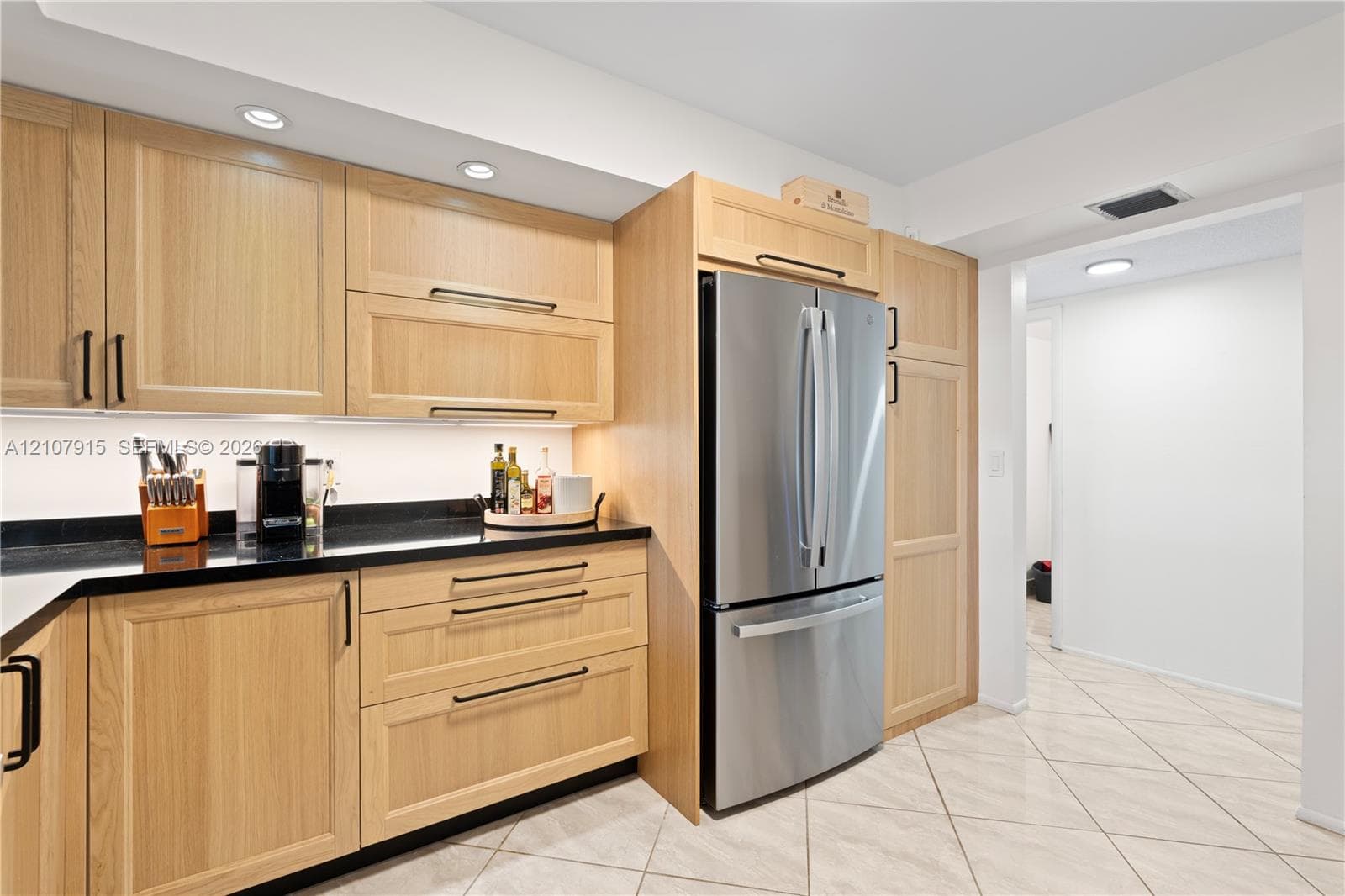 Light wood-finish cabinetry, dark stone countertops, stainless steel French-door refrigerator, under-cabinet lighting, and neutral ceramic tile flooring