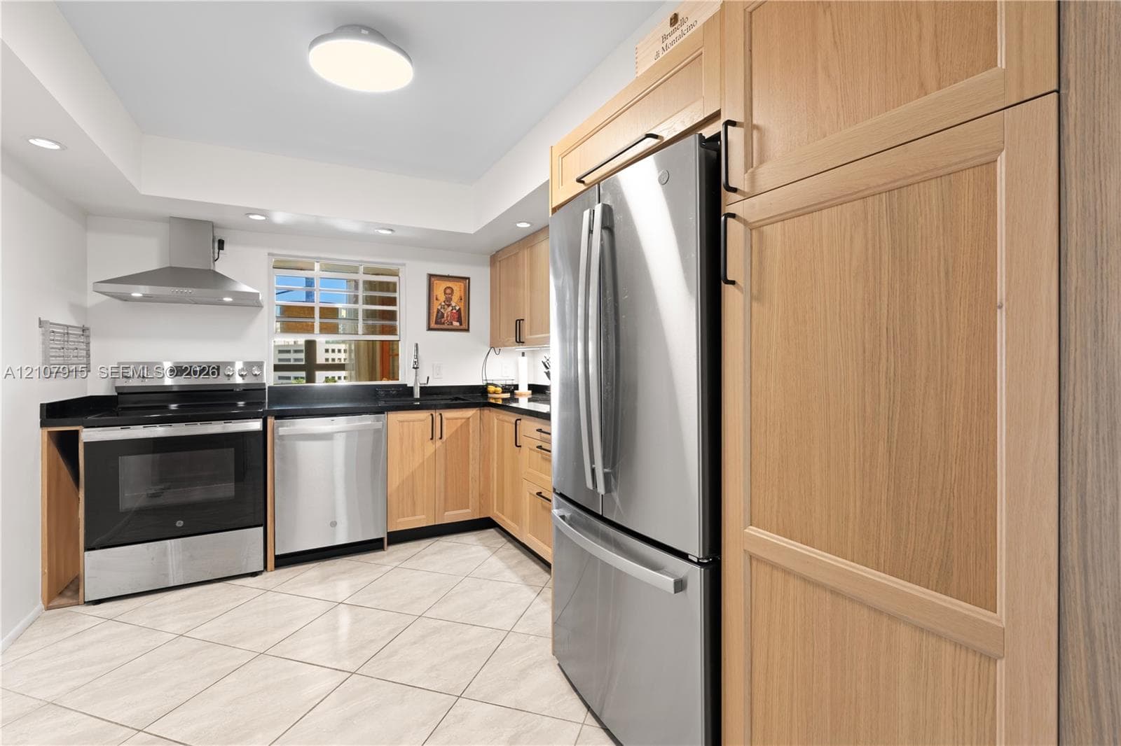 Kitchen featuring light wood-finish cabinetry, stainless steel appliances, a stainless steel range hood, dark stone countertops, and tile flooring