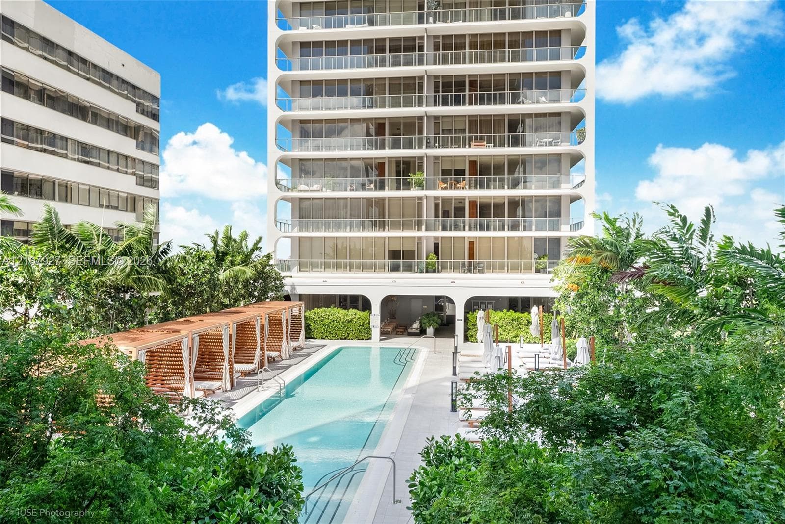 High-rise building exterior with wraparound balconies, rectangular swimming pool, integrated poolside cabanas, and mature perimeter landscaping
