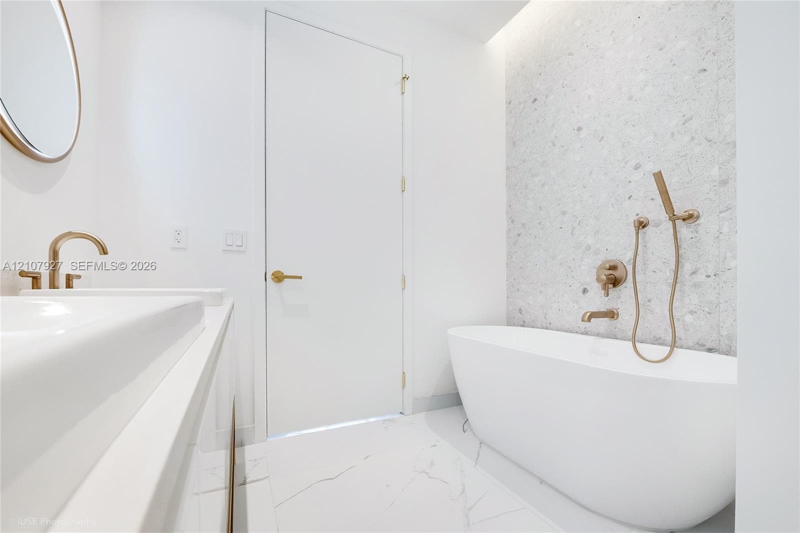 Freestanding soaking tub with wall-mounted bronze-finish fixtures, speckled stone-tiled accent wall, marble-style flooring, and integrated sink vanity with a circular mirror
