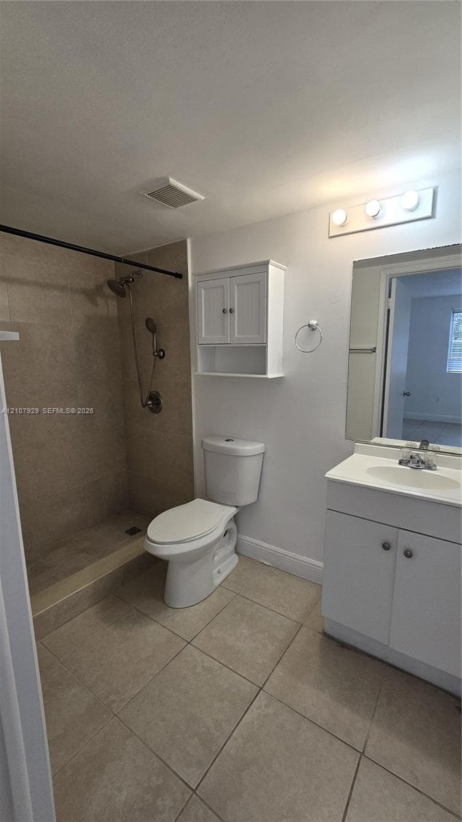 Bathroom featuring large-format neutral floor tiling, wall-mounted storage cabinetry, a multi-bulb vanity light fixture, and a tiled shower surround with adjustable shower head