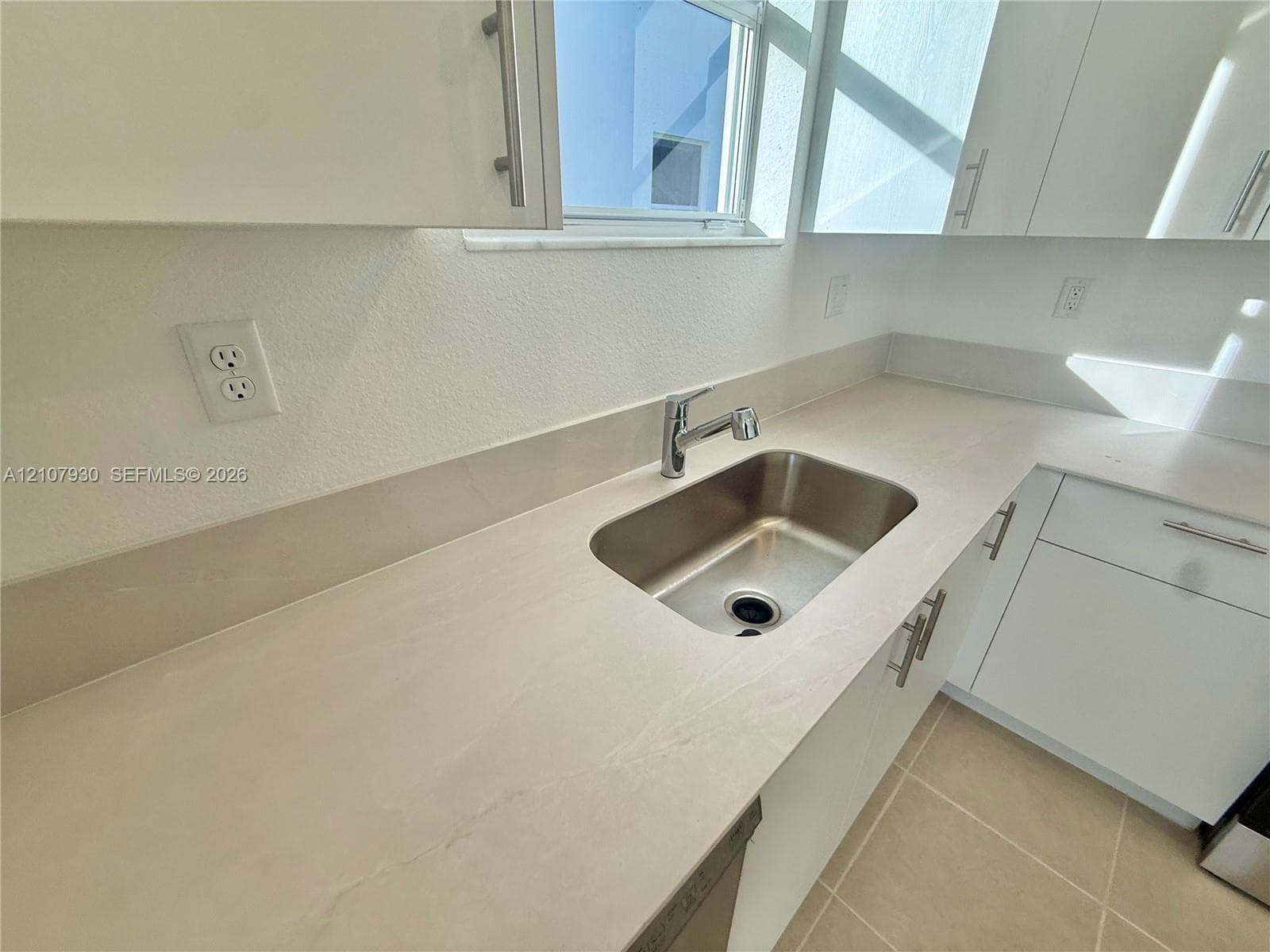 Kitchen featuring white cabinetry with brushed metal hardware, solid-surface countertops, an integrated stainless steel under-mount sink, and ceramic tile flooring