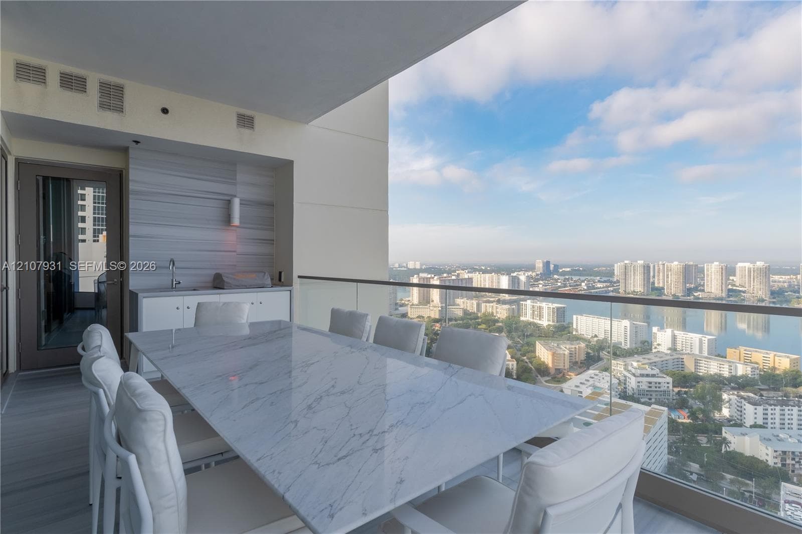 Covered balcony featuring an integrated outdoor kitchen with white cabinetry, horizontal tile wall accent, glass railing, and expansive water and city panorama