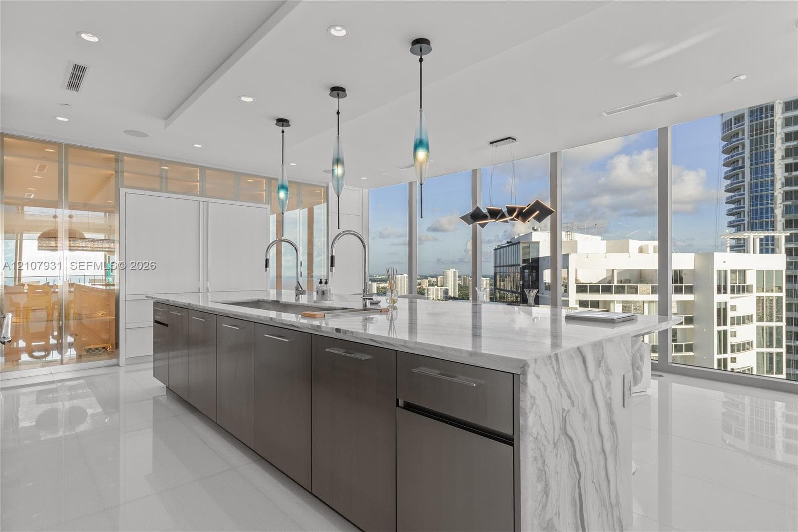 Kitchen featuring a waterfall-edge island with stone countertop, flat-panel cabinetry, dual gooseneck faucets, glass pendant lighting, and floor-to-ceiling windows