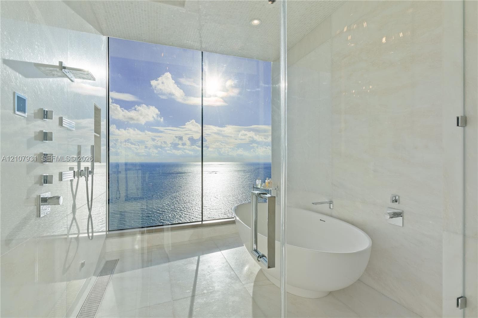 Floor-to-ceiling glass paneling, freestanding soaking tub, walk-in shower with multi-head wall-mounted shower system, and light-toned tile flooring