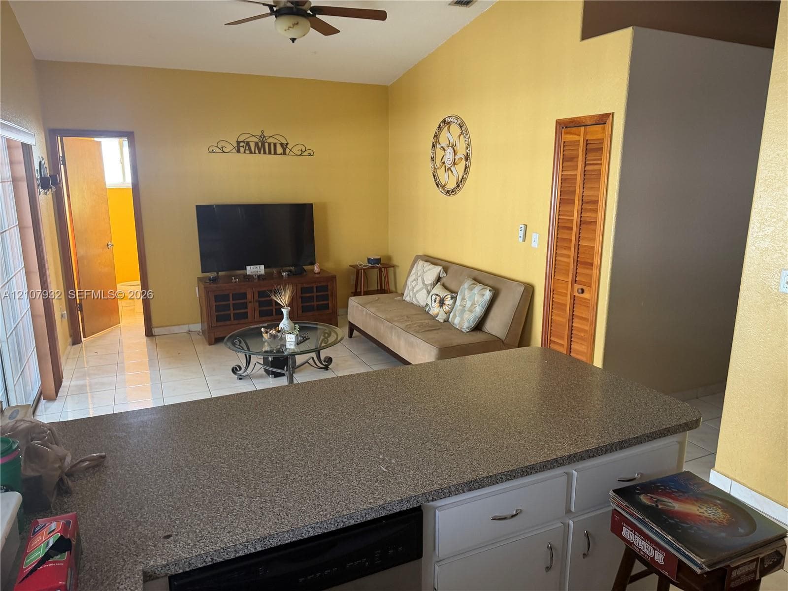 Open-concept interior featuring vaulted ceilings, neutral tile flooring, a kitchen island with speckled solid-surface countertop, and louvered closet doors