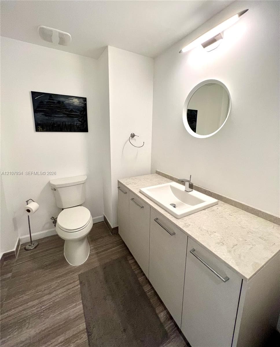 Bathroom featuring a vanity with light-toned stone countertop, integrated rectangular sink, wall-mounted circular mirror, contemporary bar-style light fixture, and wood-finish flooring