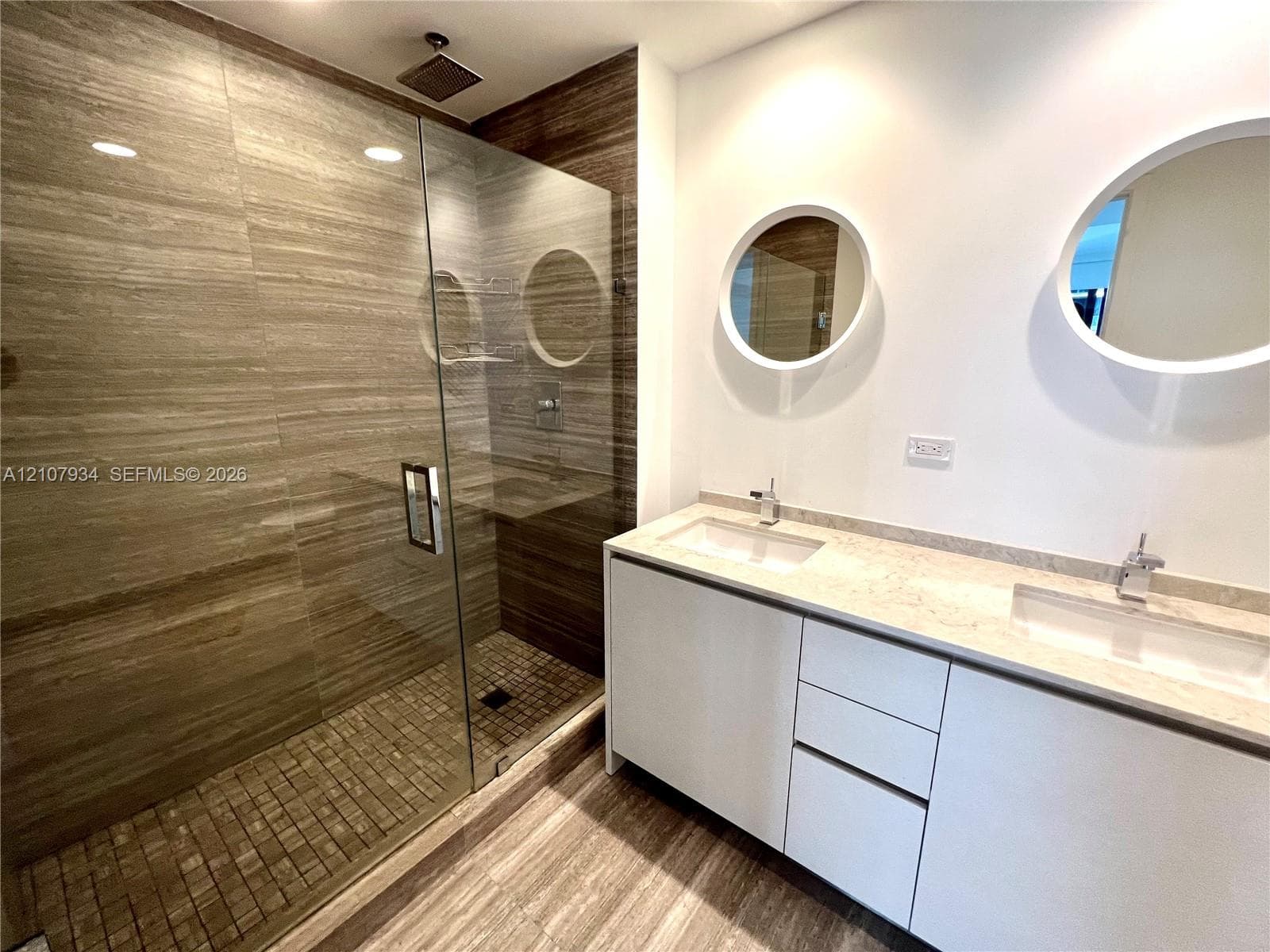Double vanity with solid surface countertop, rectangular undermount sinks, frameless glass shower enclosure, large-format tile surround, and dual circular wall-mounted mirrors