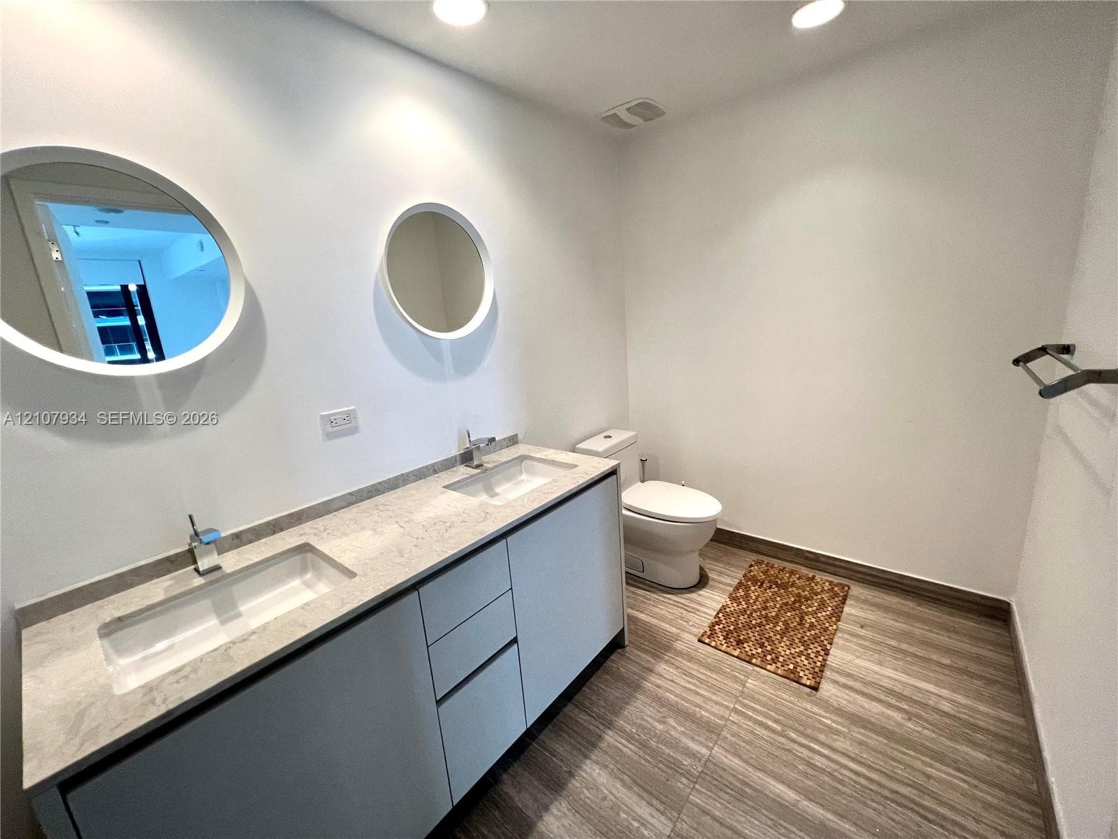 Dual-vanity cabinet featuring light-toned stone countertops, rectangular undermount sinks, chrome fixtures, two round mirrors with integrated lighting, and tile flooring