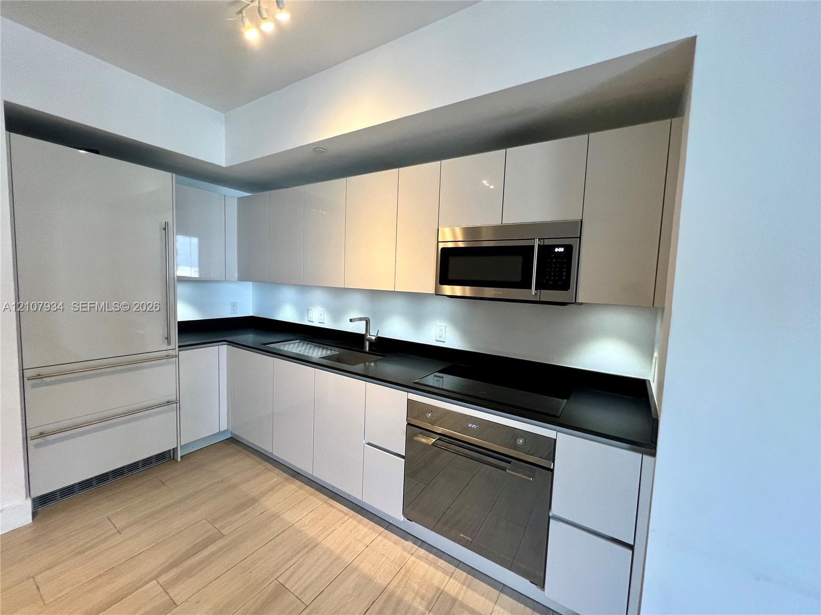 Kitchen featuring high-gloss cabinetry, dark solid-surface countertops, wood-finish flooring, and integrated stainless steel appliances including a wall oven and microwave