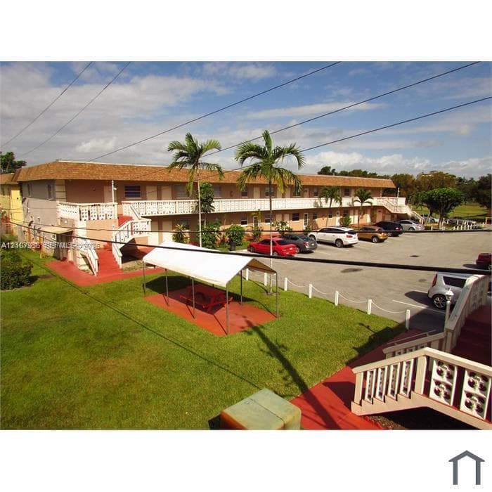 Multi-story building with a shingle-style roof, exterior staircases, designated paved parking area, and landscaped grounds featuring palm trees