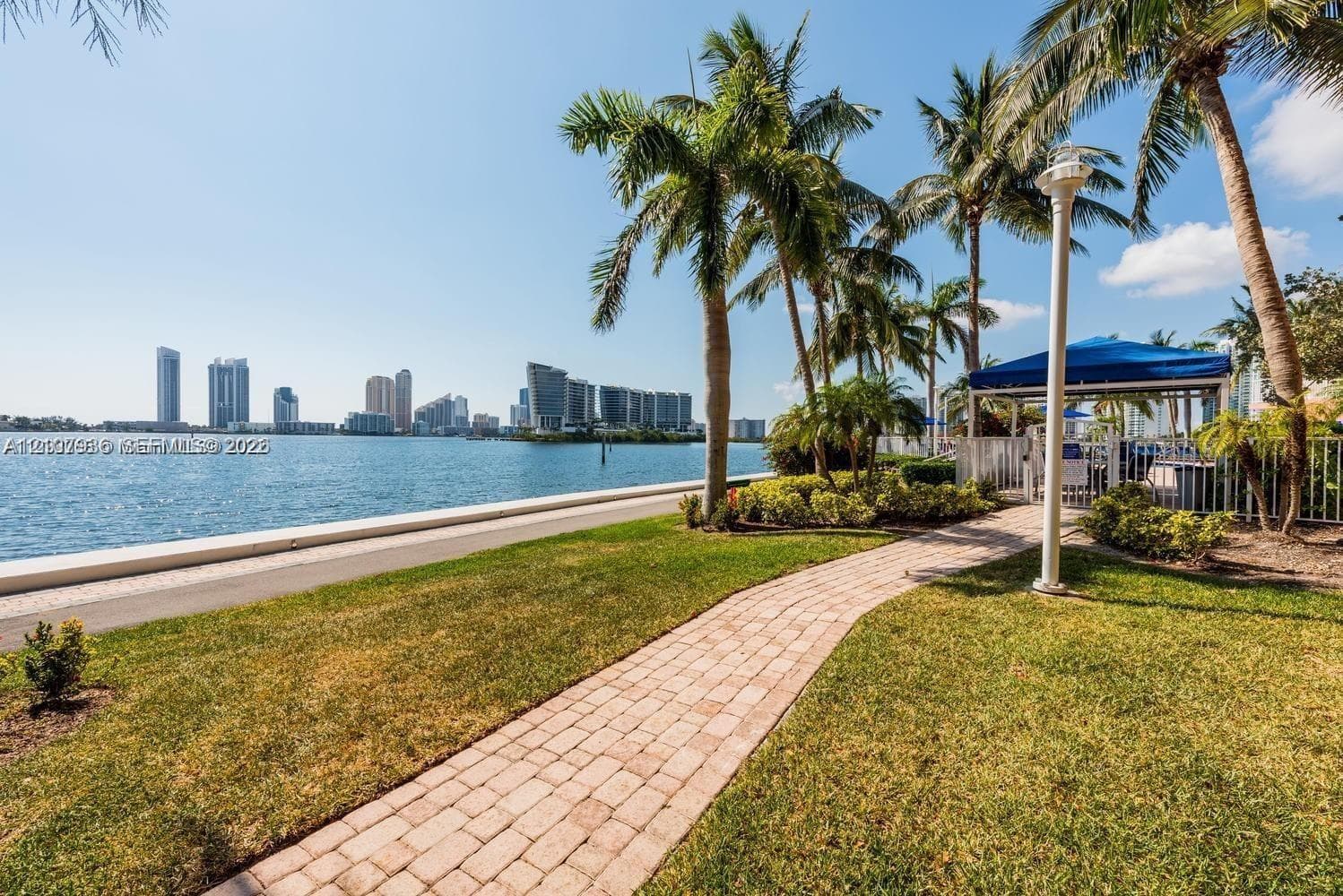 Waterfront grounds featuring a brick paver pathway, mature palm trees, coastal landscaping, and perimeter fencing