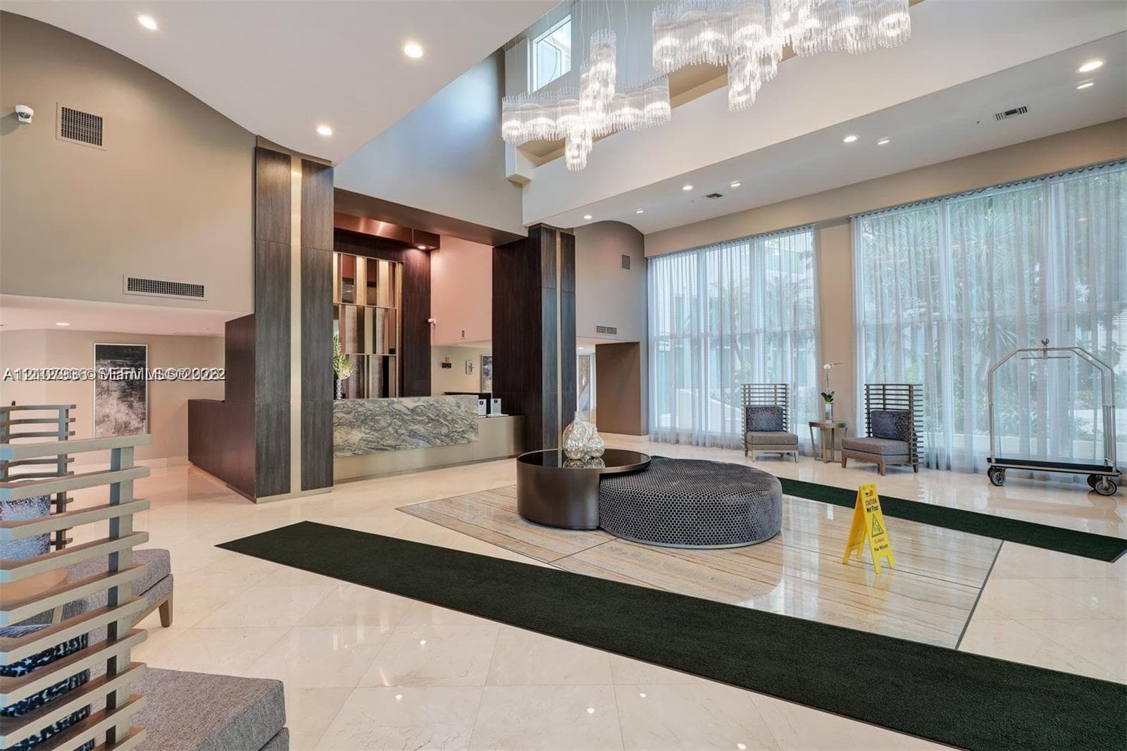 Double-height ceiling lobby featuring stone-finish flooring, a tiered crystal chandelier, floor-to-ceiling windows, and a reception desk with stone-finish facade