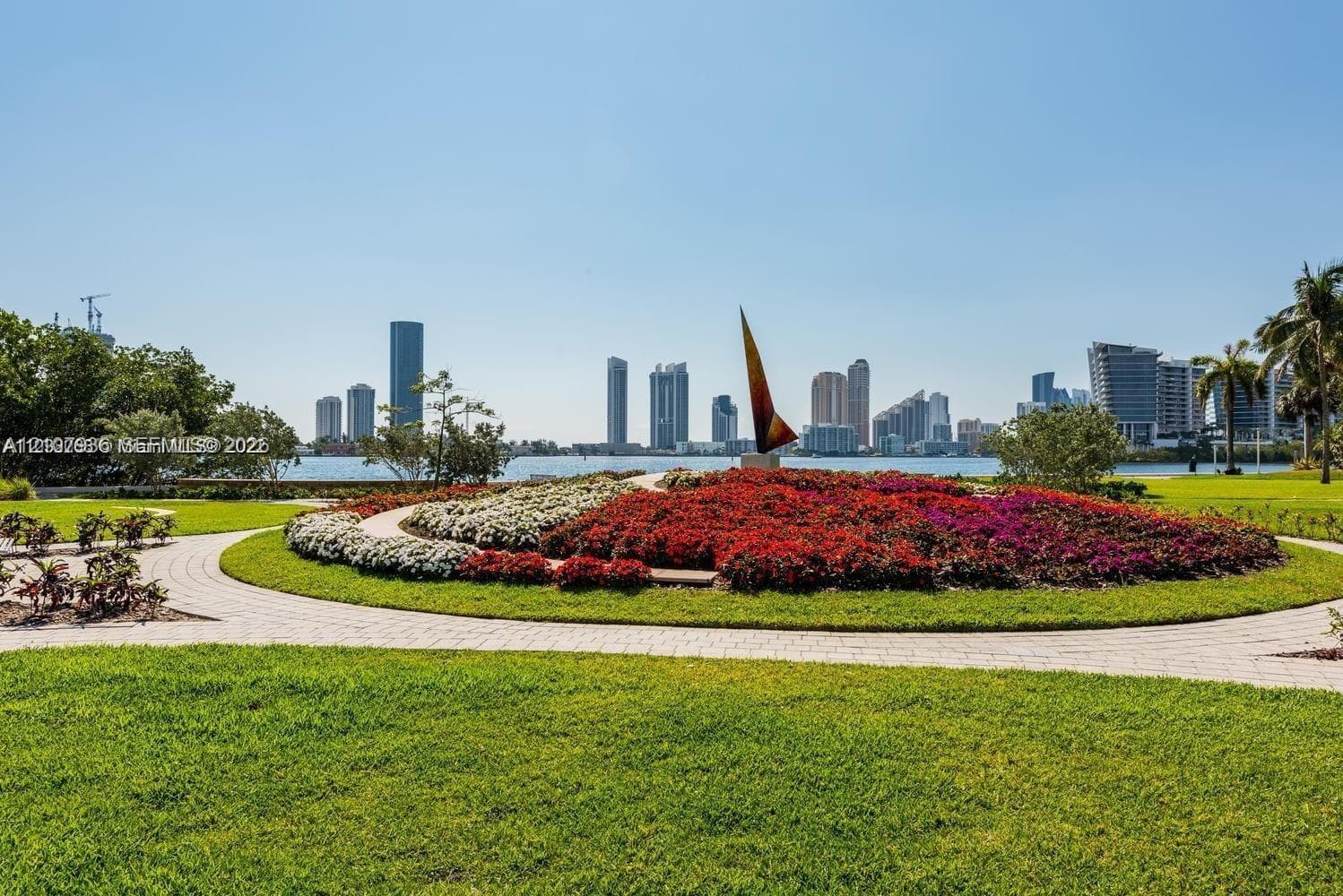 Manicured lawn featuring a circular stone paver path, expansive floral landscaping, decorative sculpture, and water frontage with a cityscape backdrop
