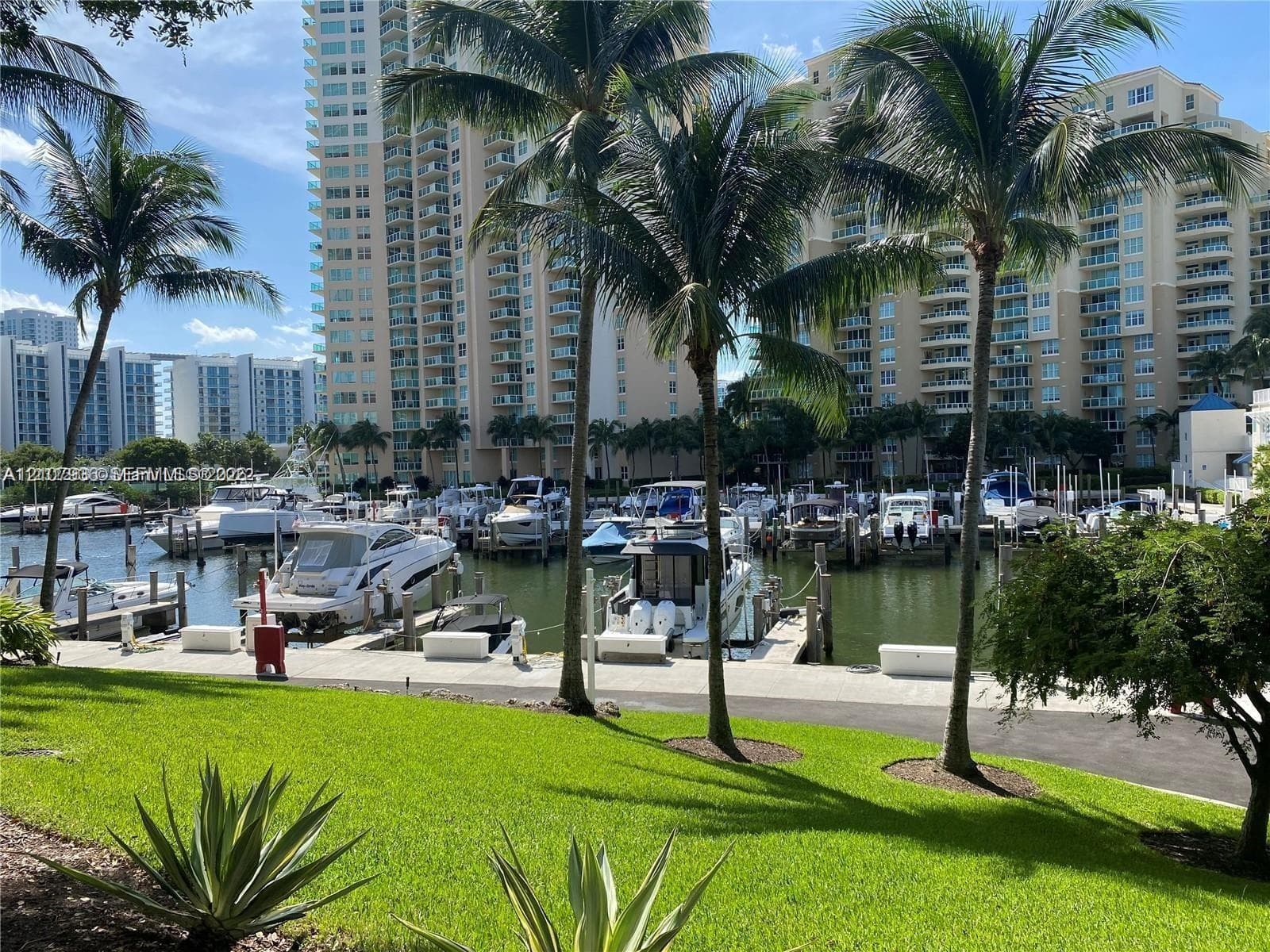 Marina featuring multiple boat slips, paved waterfront walkway, and manicured green space with mature palm trees