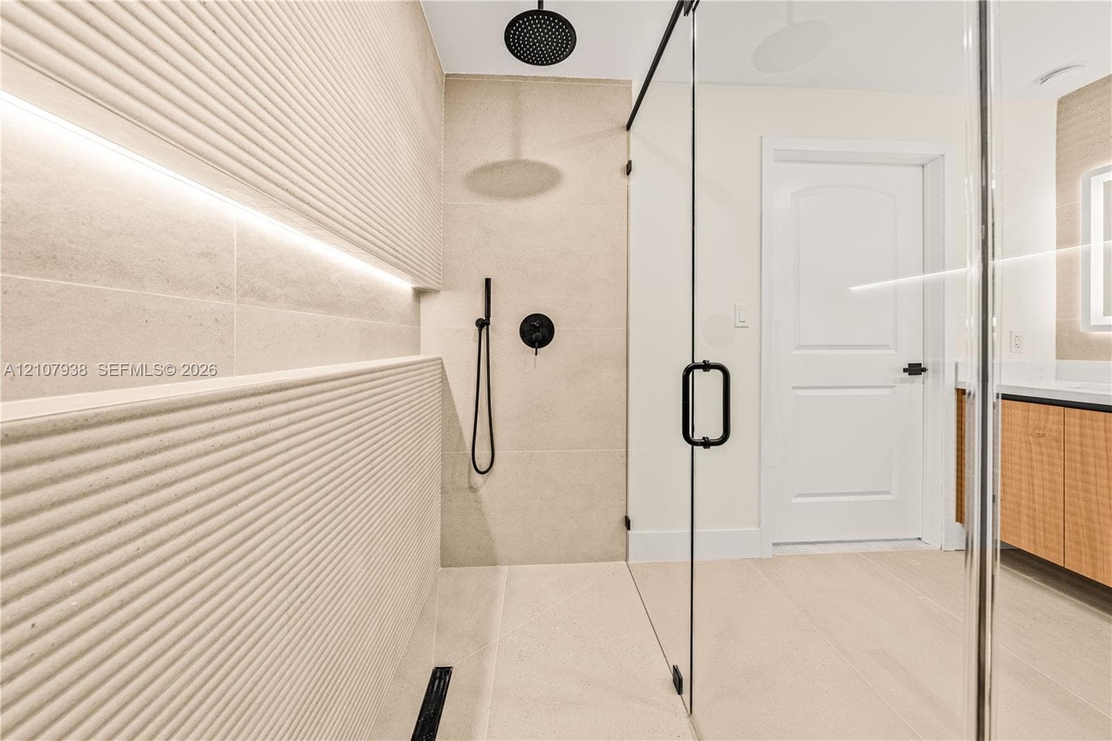 Walk-in shower featuring a frameless glass enclosure, matte black rainfall shower head with handheld attachment, integrated wall niche lighting, and textural linear-patterned tile accents