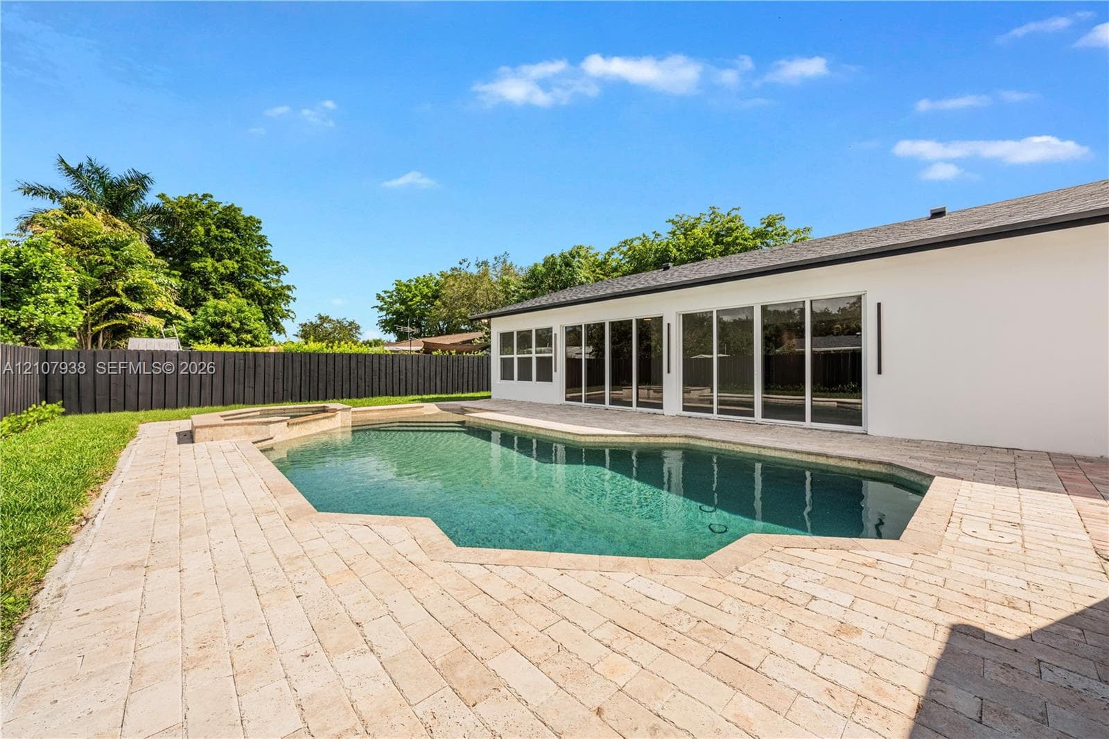 Custom-shaped in-ground pool featuring a built-in spa, expansive stone paver deck, white exterior wall, and floor-to-ceiling glass sliding doors