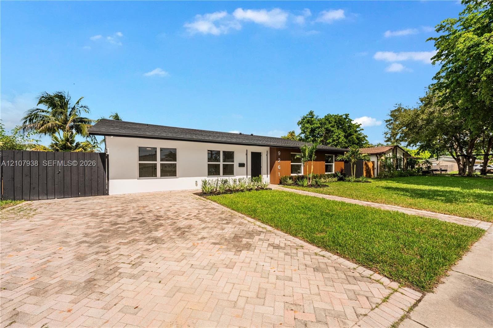 Single-story exterior featuring white stucco and wood-panel siding, a herringbone-patterned paver driveway, a black horizontal-slat privacy fence, and a dark shingled roof