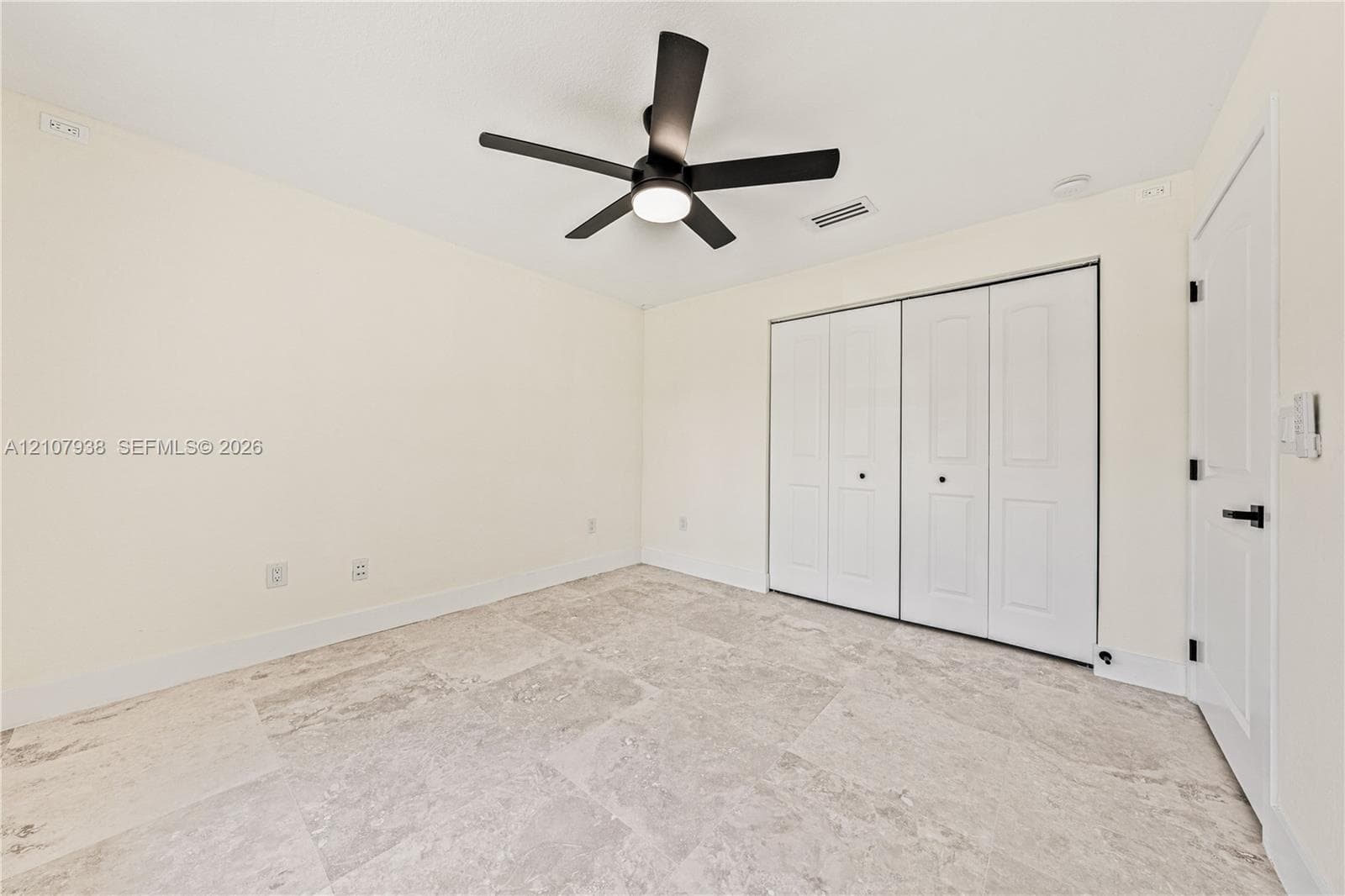 Room featuring neutral-toned stone tile flooring, a matte black multi-blade ceiling fan with integrated lighting, and a multi-panel closet with matte black hardware