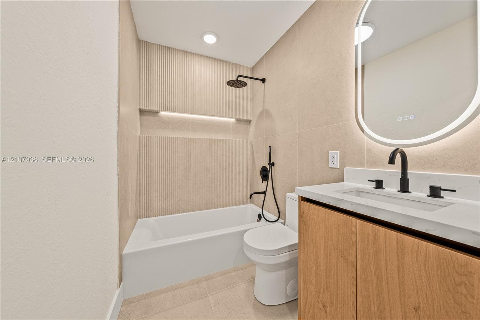 Modern bathroom featuring a wood-finish vanity with a stone countertop, an oval LED-backlit mirror, large-format neutral wall tile, and a built-in recessed shower niche with integrated lighting