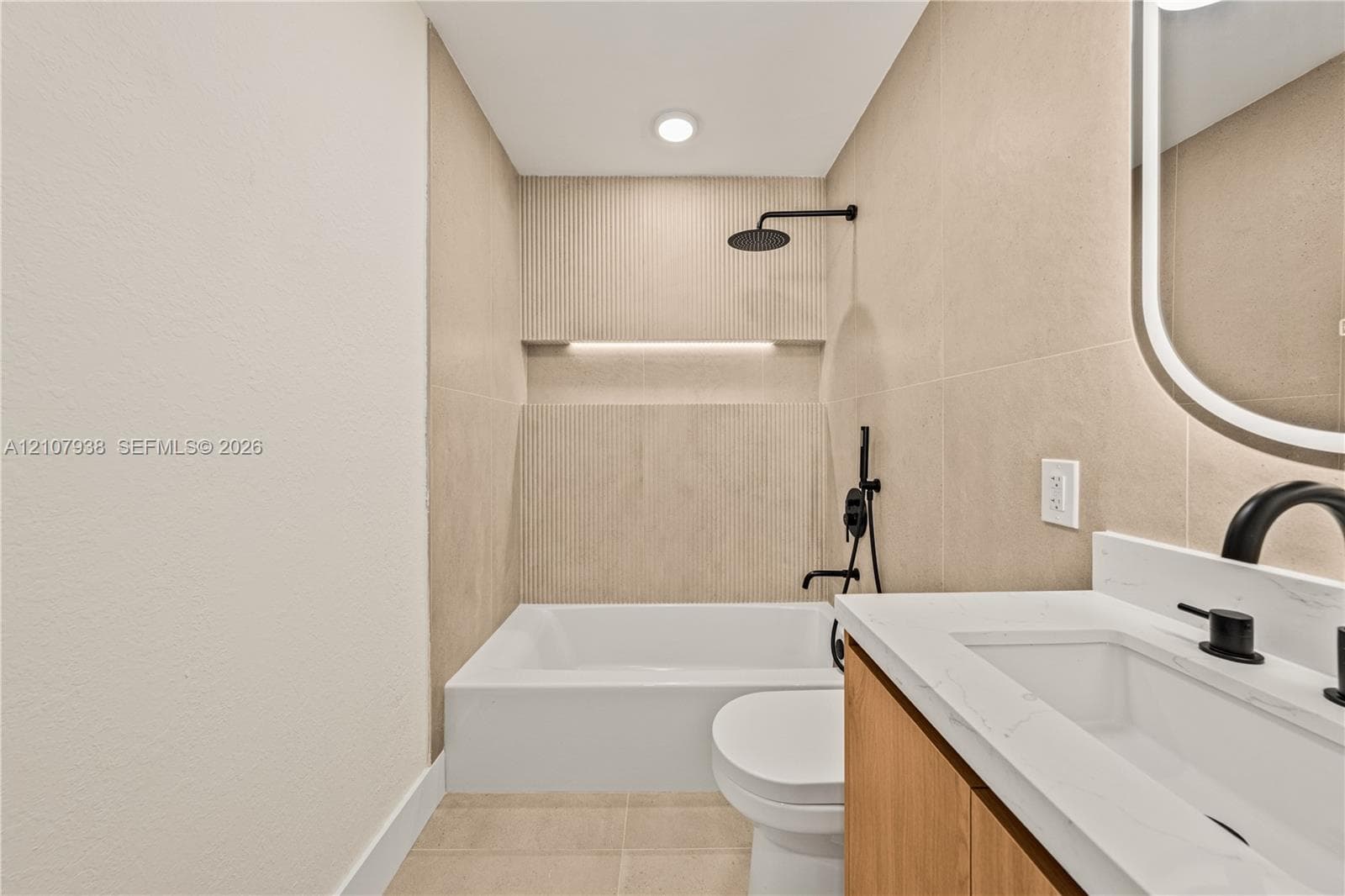 Bathroom featuring a vanity with a light wood-finish cabinet, stone-surface countertop, and matte black fixtures, paired with a shower-tub combination featuring recessed wall niche lighting and floor-to-ceiling tile work