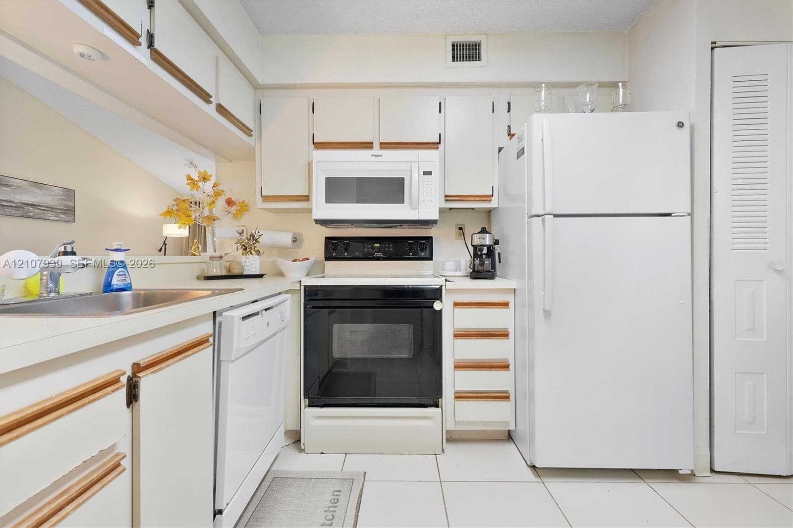 Kitchen featuring white cabinetry with wood-finish trim, white appliances including a built-in microwave and dishwasher, a stainless steel sink, and ceramic tile flooring