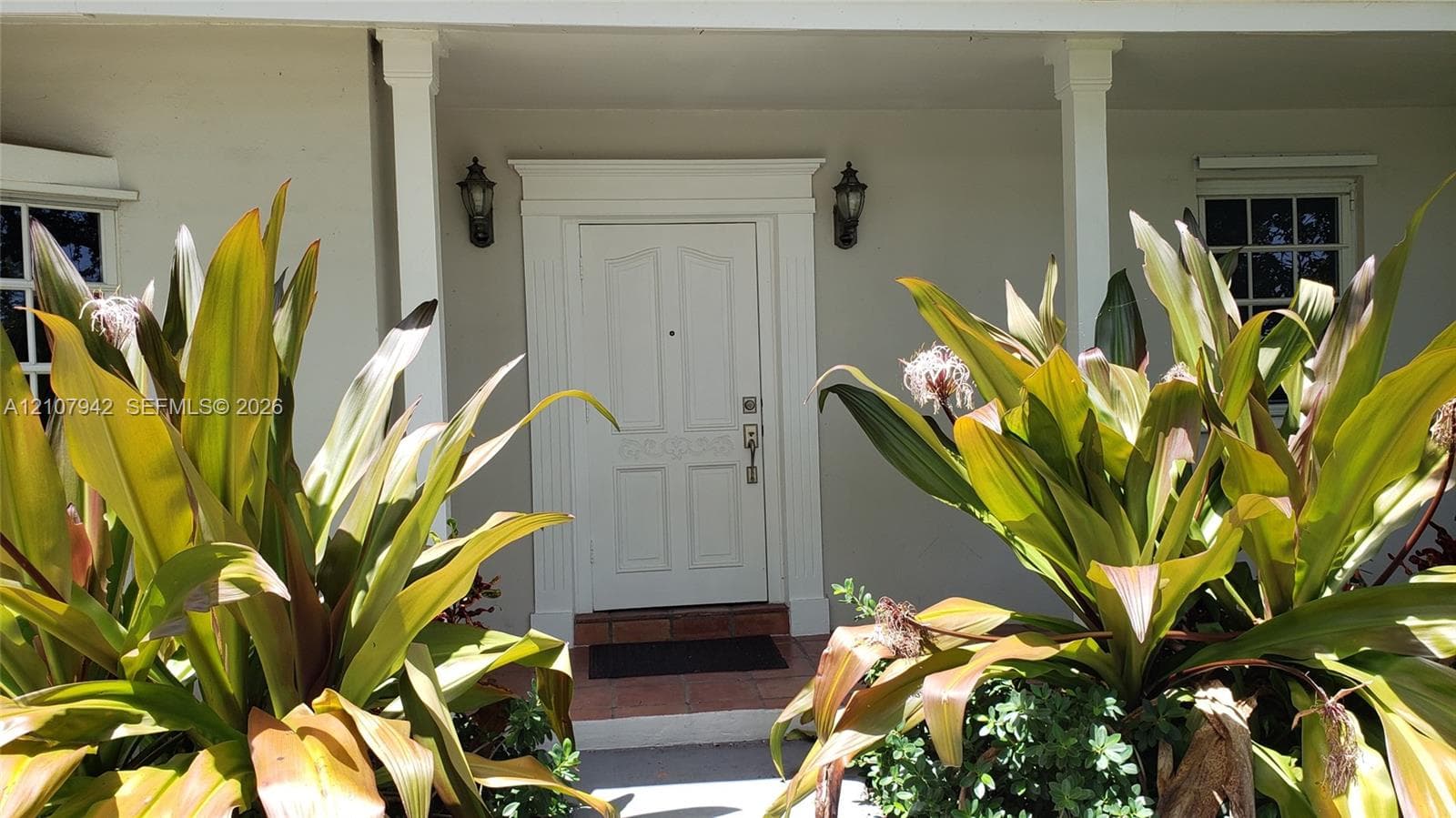 Covered front porch featuring a paneled entry door with architectural trim, dual wall-mounted exterior fixtures, and tiled flooring