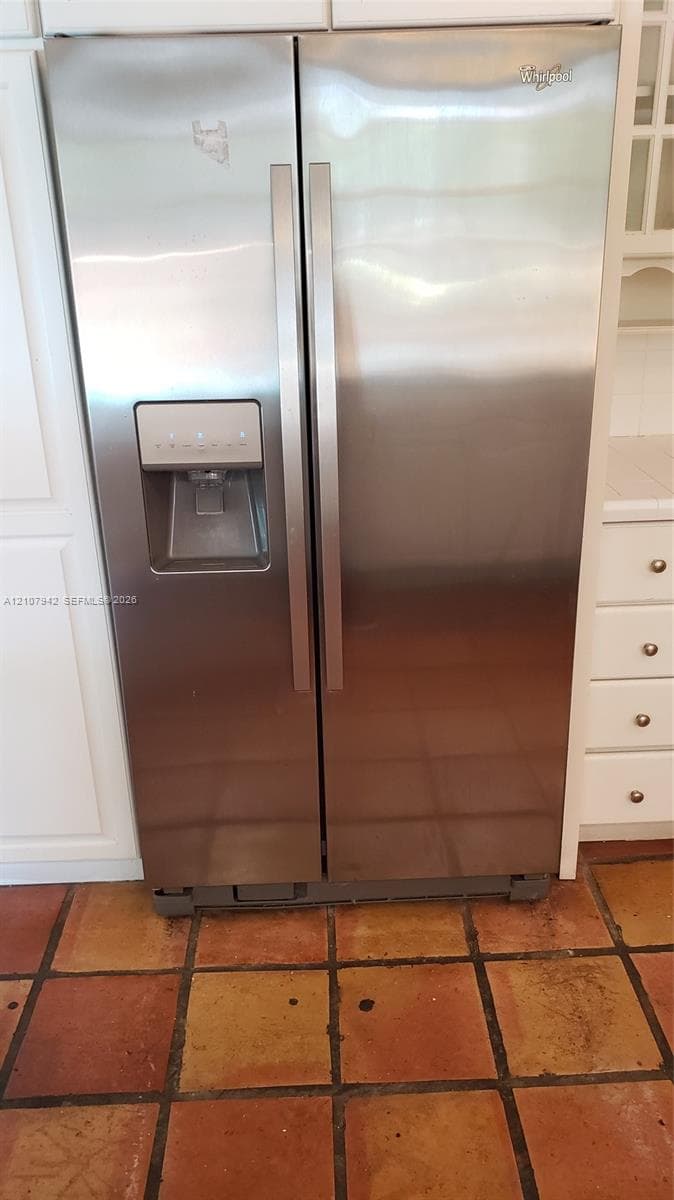 Stainless steel side-by-side refrigerator featuring an integrated water and ice dispenser, surrounded by white cabinetry and finished with terracotta tile flooring
