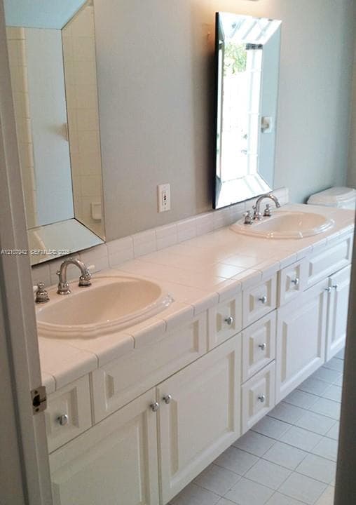 Double vanity featuring white cabinetry, a tiled countertop, dual drop-in sinks with metal fixtures, and rectangular wall-mounted mirrors